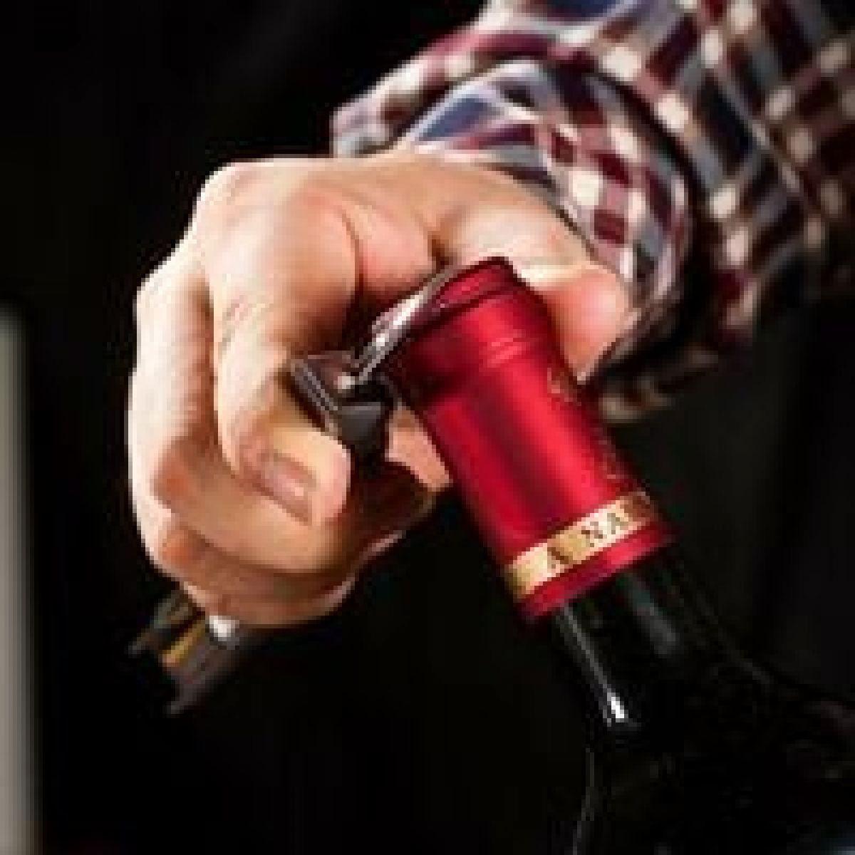 Eine Hand öffnet eine Weinflasche mit einem Korkenzieher. Die Flasche hat einen roten Deckel. Die Person trägt ein kariertes Hemd.