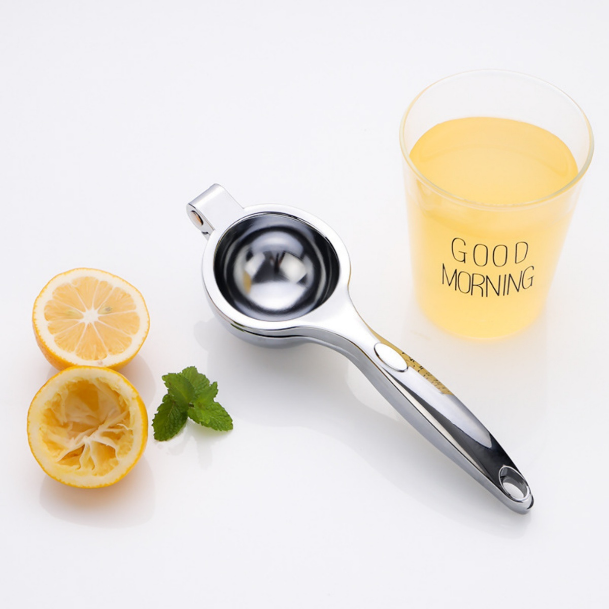 Eine Zitronenpresse mit halben Zitronen, Minze und einem Saftglas mit der Aufschrift "Good Morning" auf weißer Oberfläche.