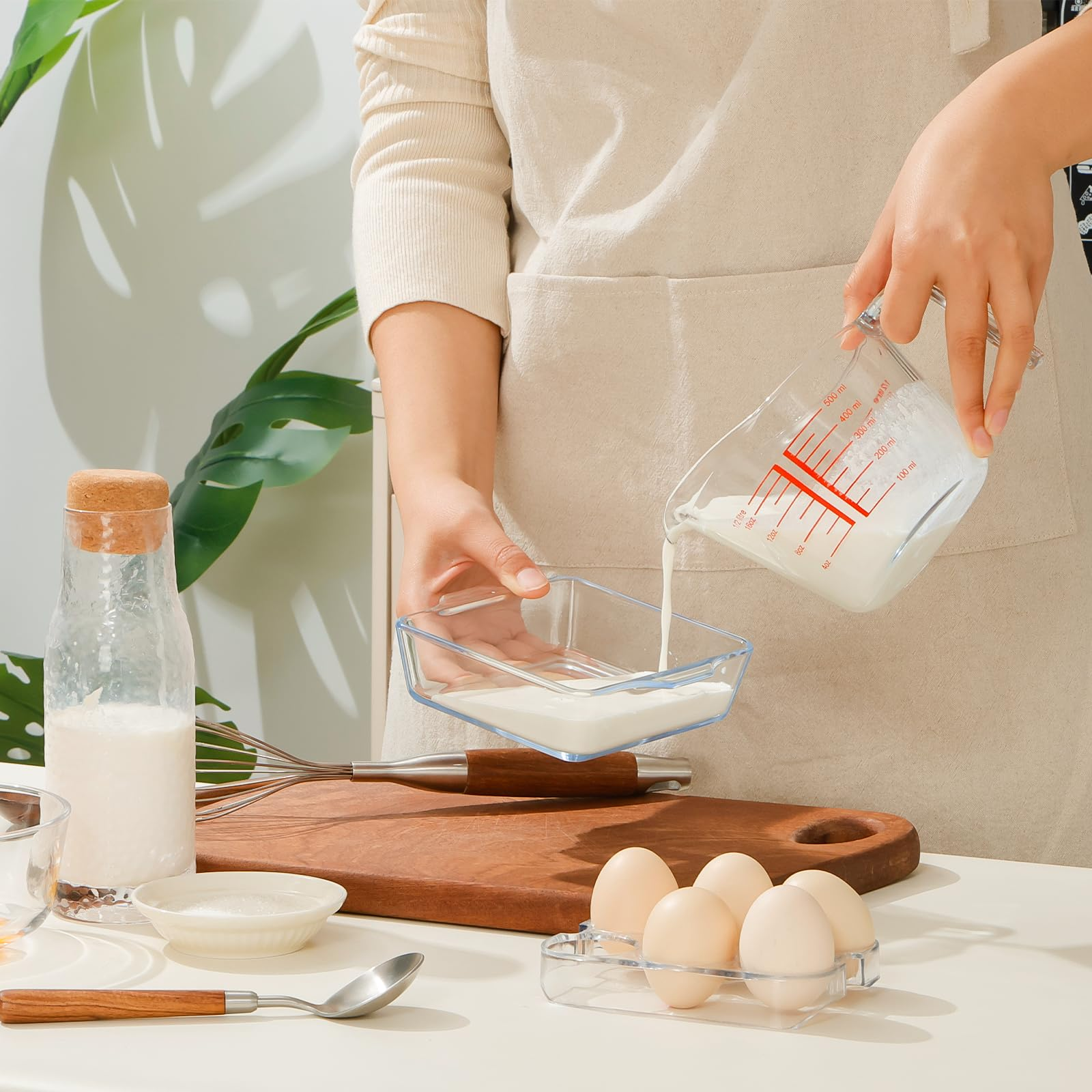 Eine Person gießt Milch aus einem Messbecher in eine Glasschale. Eier, Schneebesen und weitere Zutaten sind sichtbar.