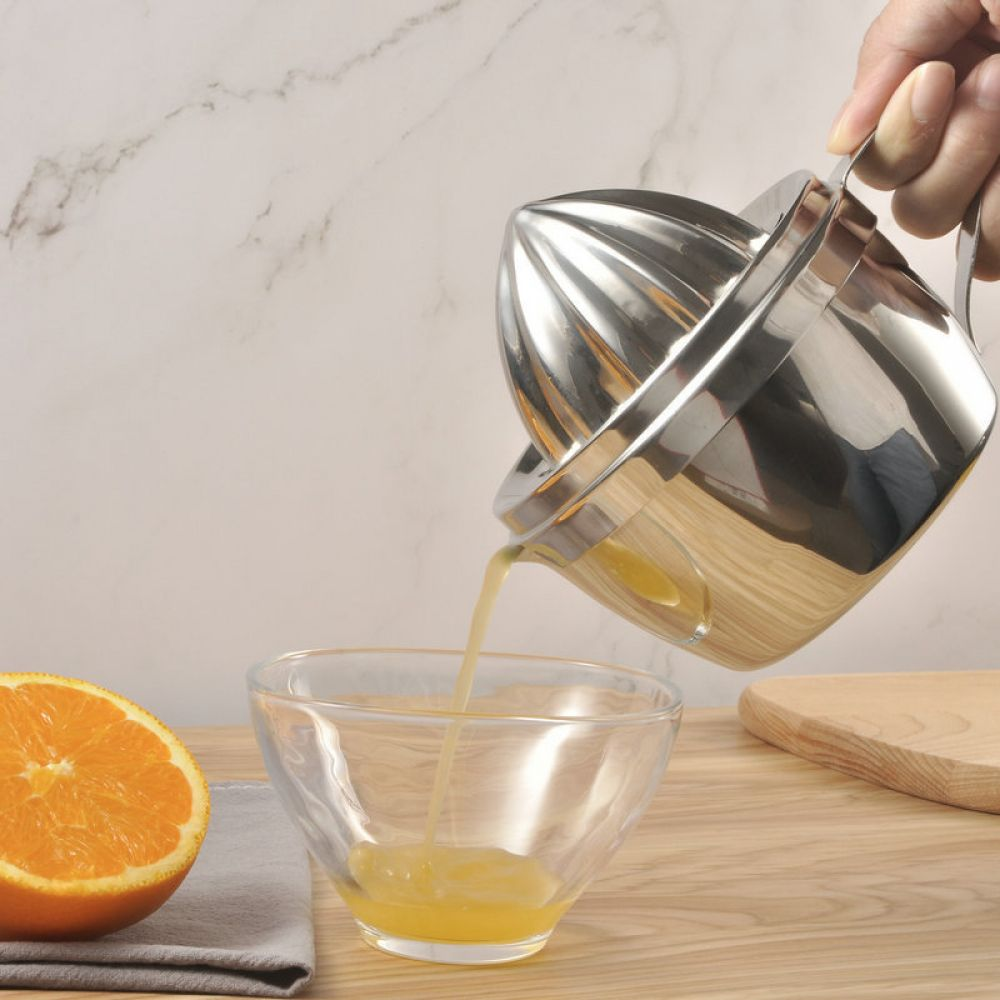 Eine Hand presst Orangensaft in eine Glasschüssel. Eine halbe Orange liegt neben einem grauen Tuch. Im Hintergrund befindet sich ein Schneidebrett.