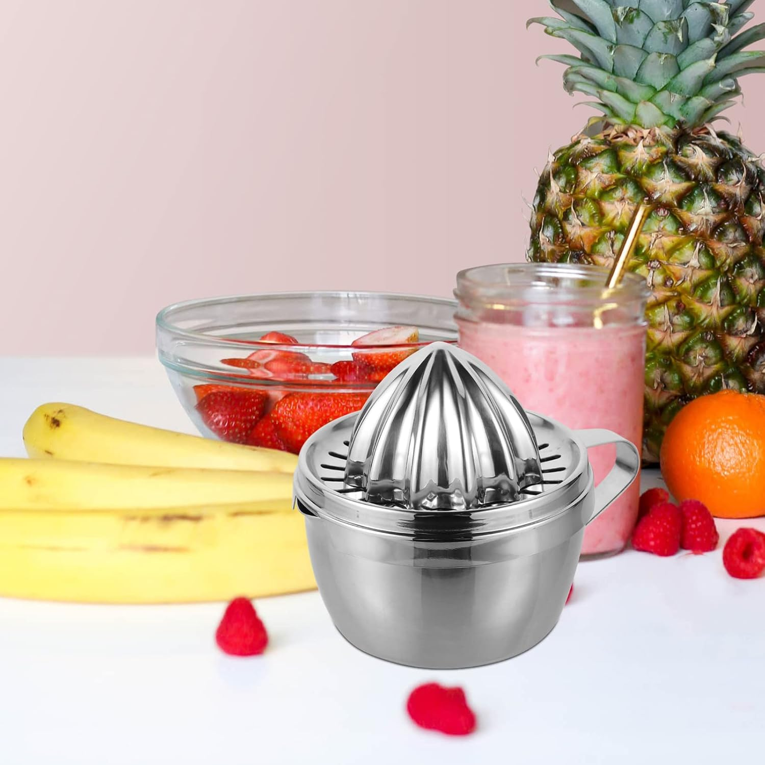 Ein Metall-Entsafter steht neben einem Glas rosa Smoothie, Früchten und einer Ananas. Bananen und Himbeeren sind ebenfalls sichtbar.