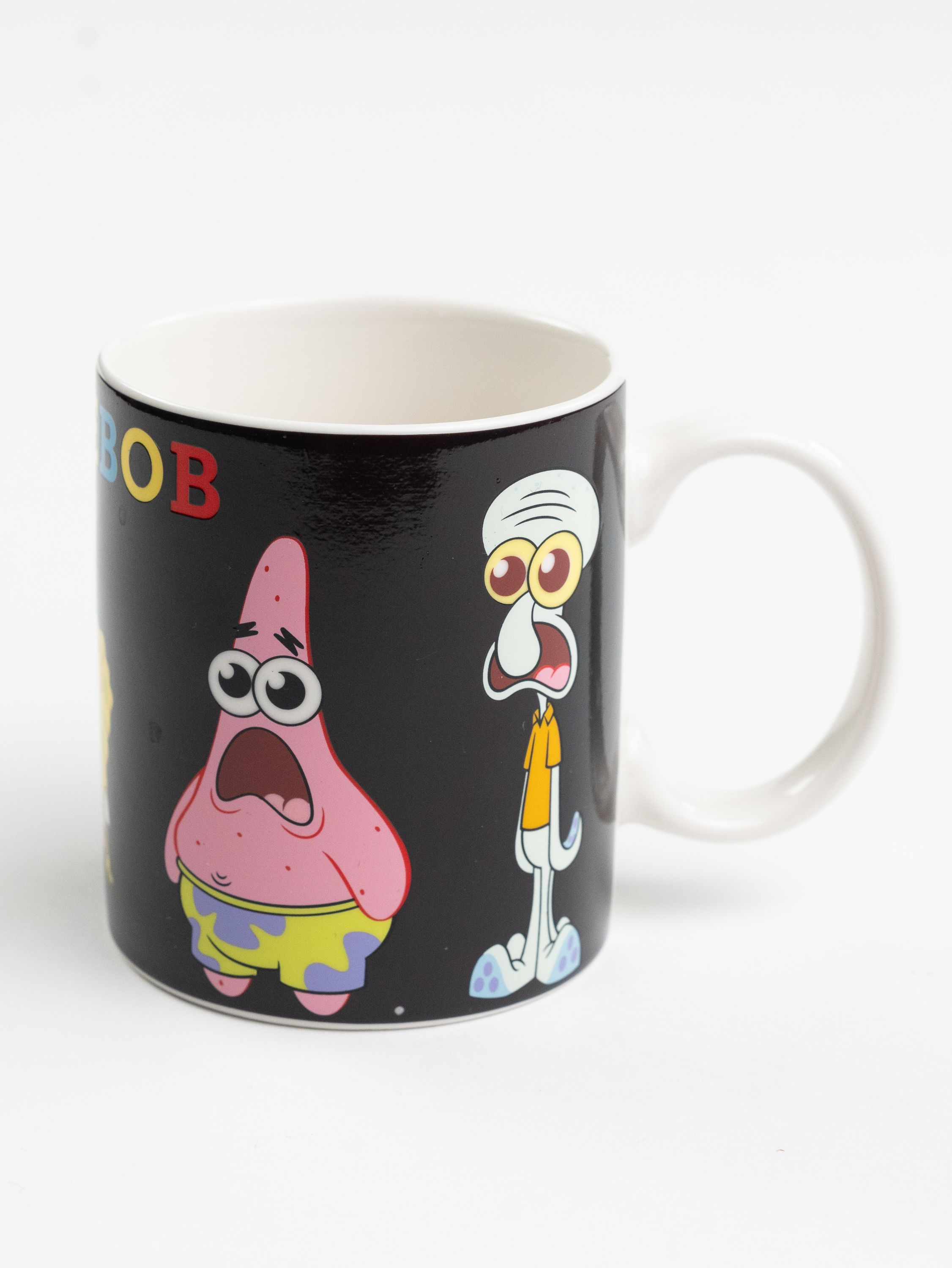 Eine Tasse mit SpongeBob-Figuren Patrick und Thaddäus auf schwarzem Hintergrund. Weißer Griff und Innenraum.