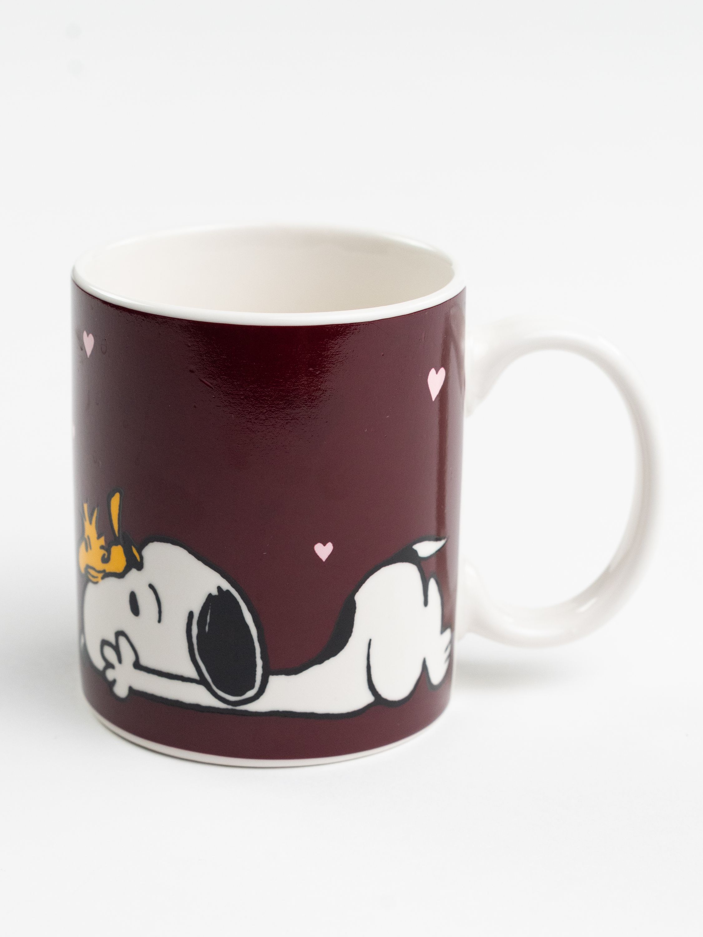 Eine Tasse mit Snoopy darauf, bordeauxfarben. Sie hat einen weißen Griff und eine weiße Innenseite. Herzen sind auf der Tasse.
