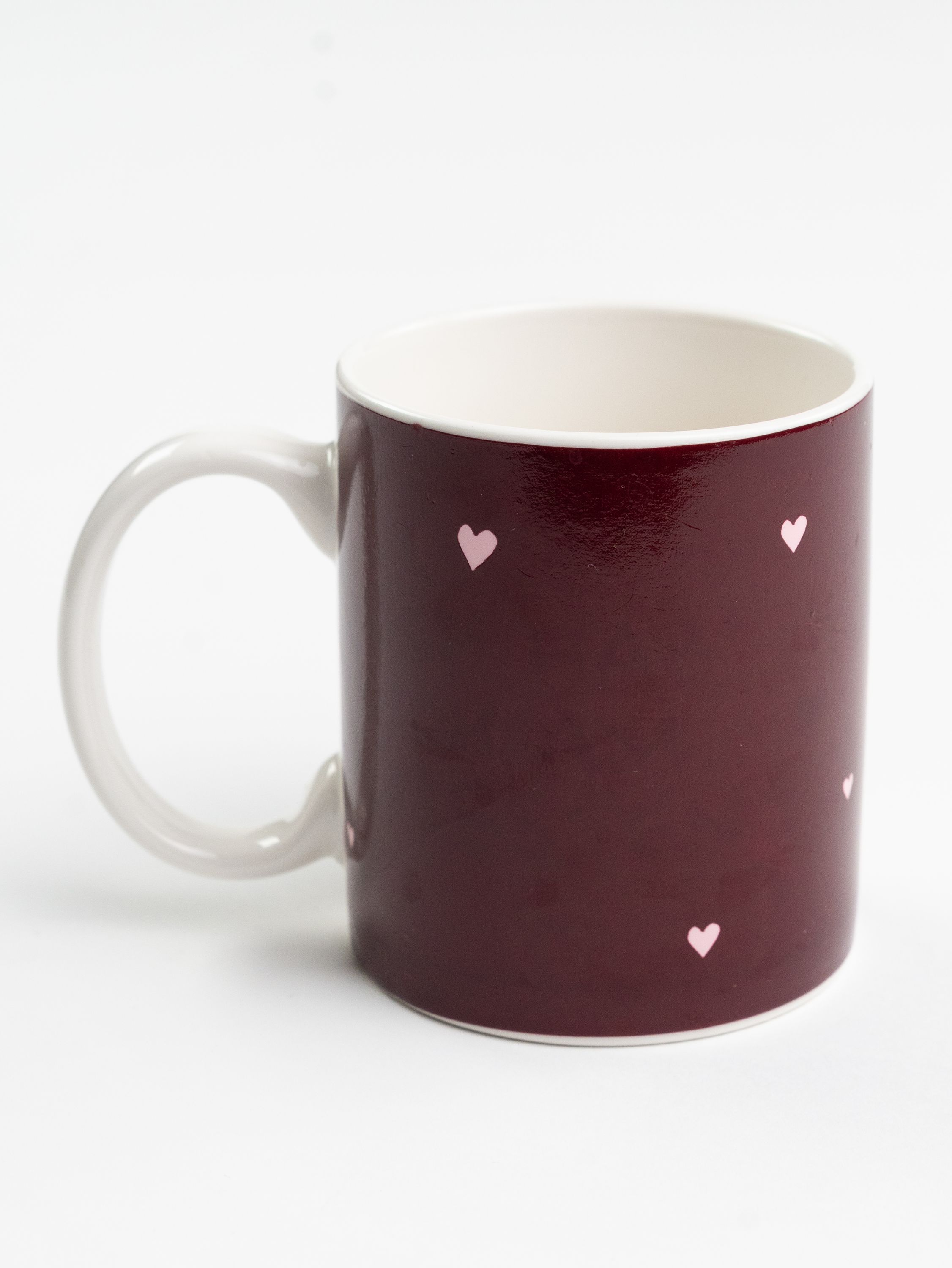 Eine weinrote Tasse mit rosa Herzen. Sie hat einen weißen Henkel und ein weißes Inneres, vor weißem Hintergrund.