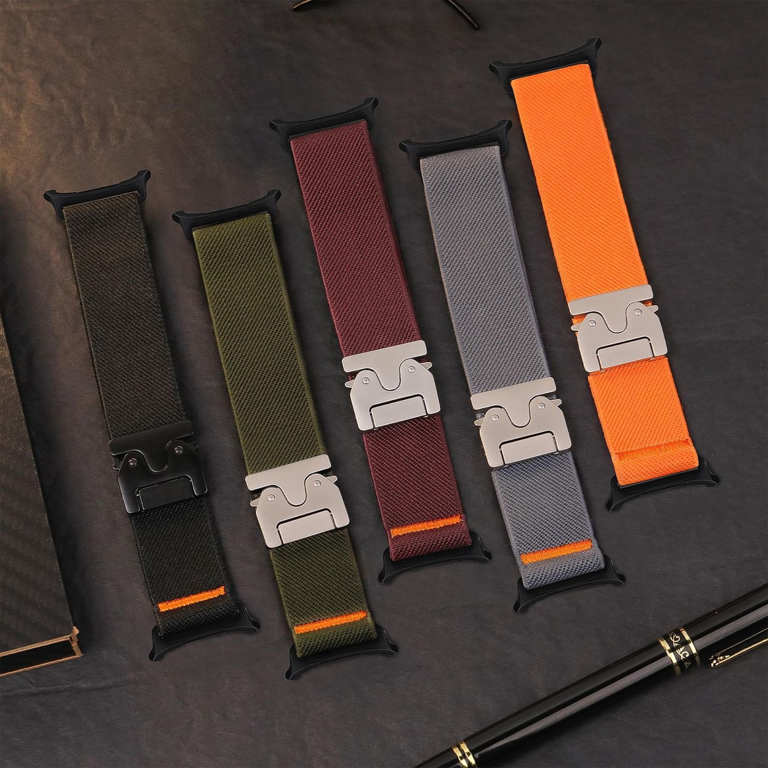 Cinque cinturini per orologi di diversi colori: nero, verde, bordeaux, grigio e arancione. Sono disposti su una superficie scura.