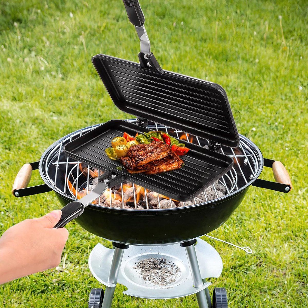 Ein Grill mit Essen darauf, gehalten von einer Hand. Der Grill steht über glühenden Kohlen, im Hintergrund Gras.