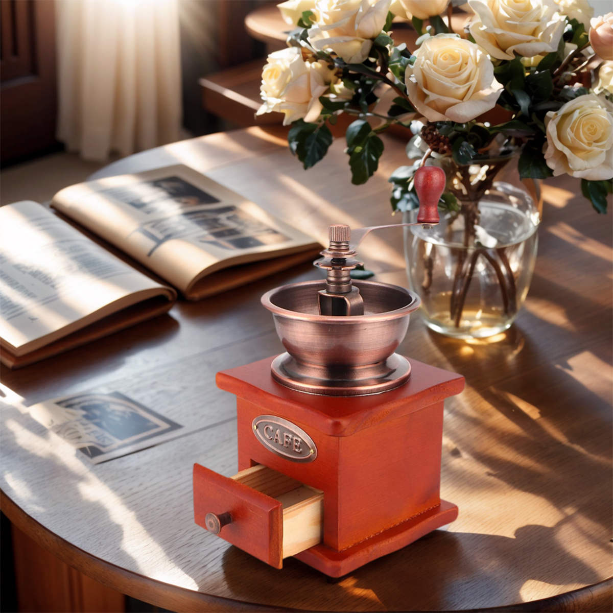 Eine hölzerne Kaffeemühle mit offener Schublade, ein Buch und eine Vase mit Rosen auf einem Holztisch.