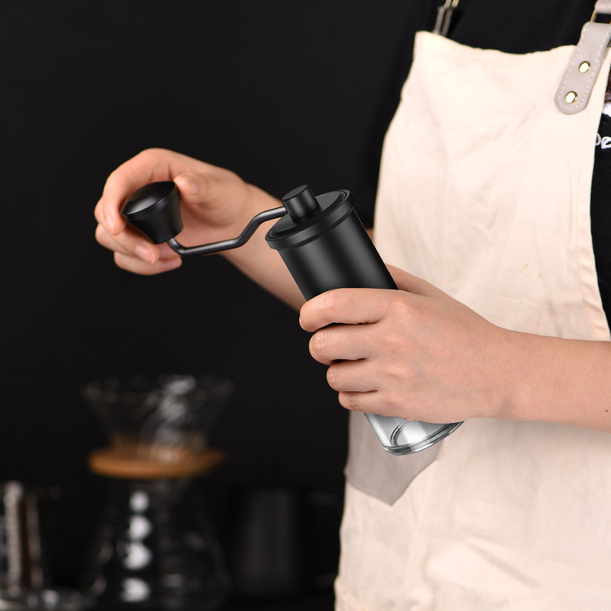 Eine Person hält eine schwarze manuelle Kaffeemühle. Eine weiße Schürze wird getragen. Im Hintergrund ist Kaffeezubereitungszubehör.