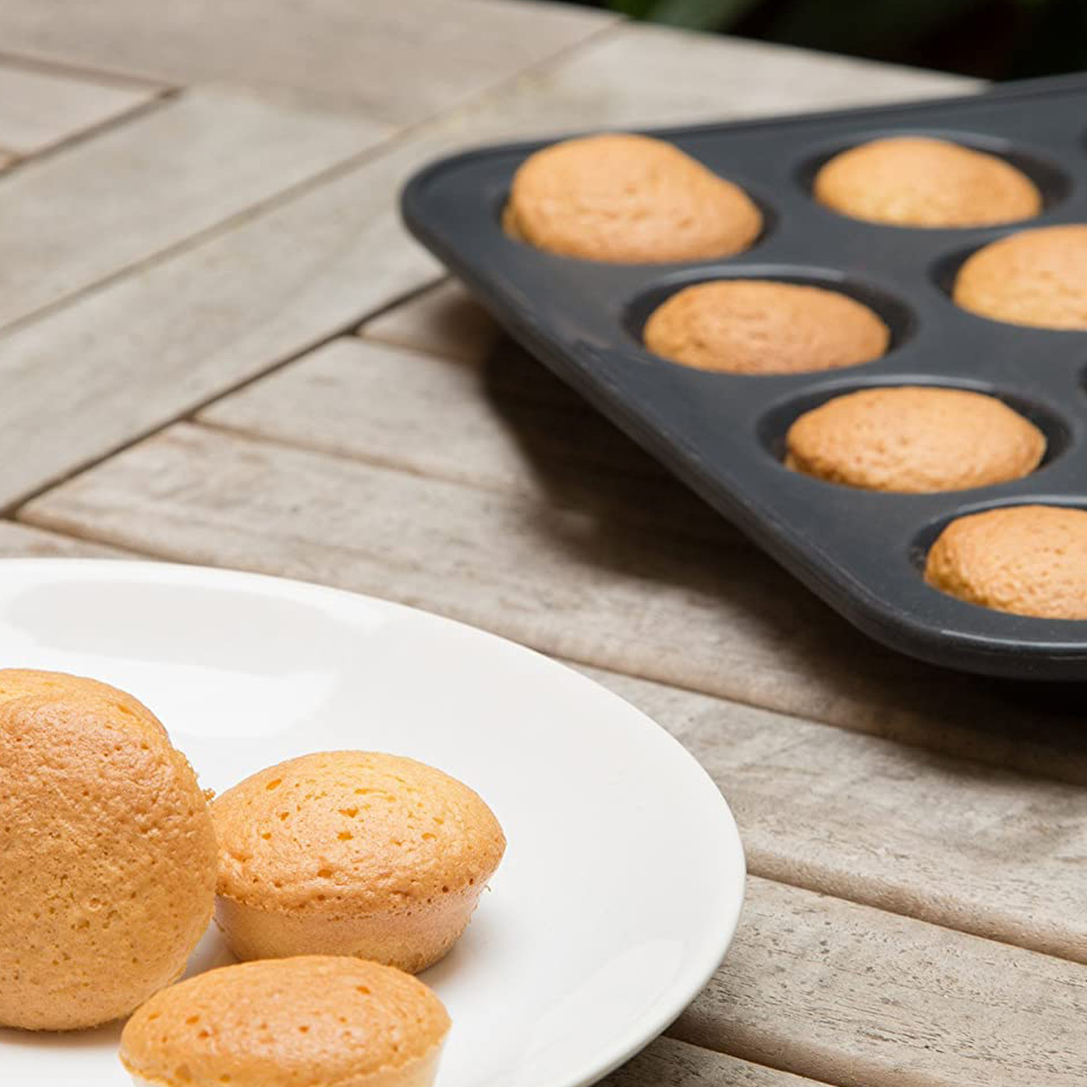 Mehrere goldene Muffins auf einem weißen Teller und in einer Muffinform auf einem Holztisch. Im Hintergrund sieht man grünes Laub.