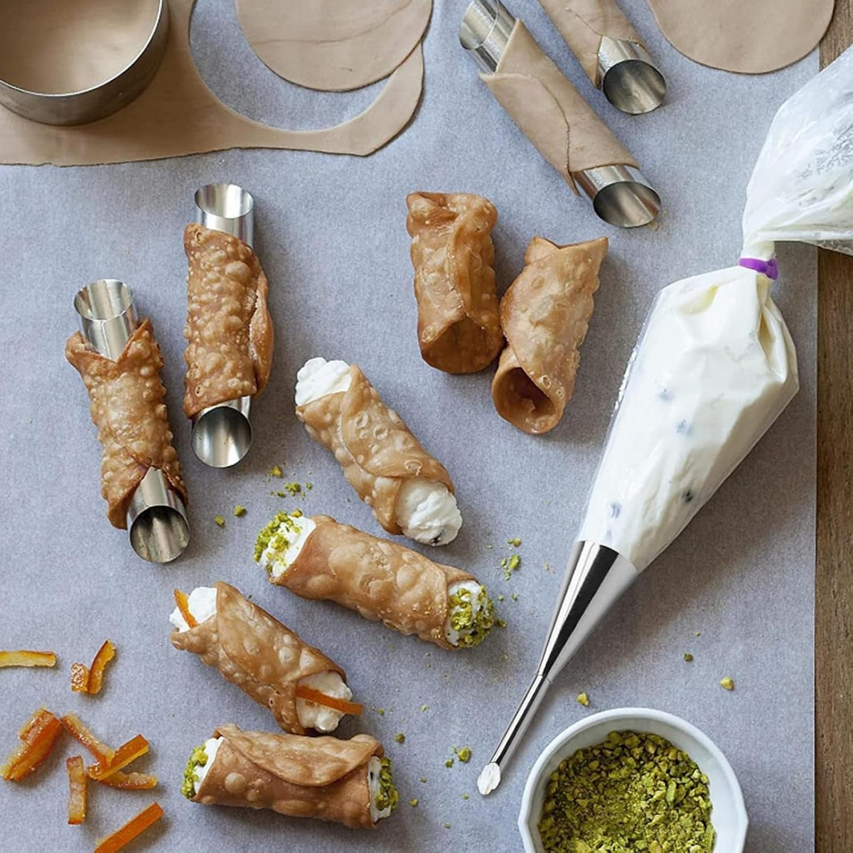 Cannoli auf Pergamentpapier mit cremefarbenen Backbeuteln, Pistazien und Orangenschalen. Einige Cannoli befinden sich auf Metallzylindern.
