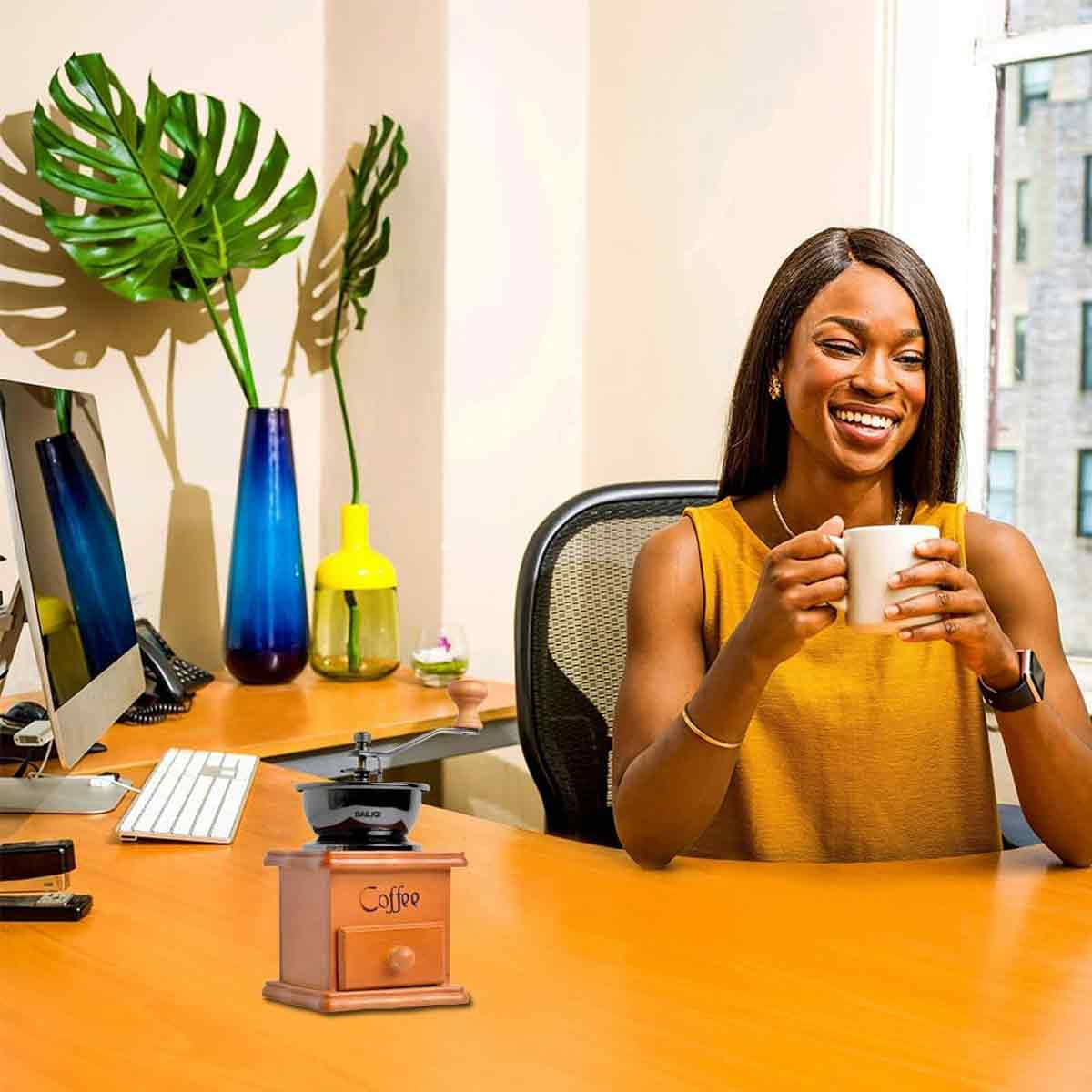 Frau in gelbem Oberteil lächelt, hält eine Tasse. Kaffeemühle auf dem Schreibtisch mit Monitor und Vasen. Fenster im Hintergrund.