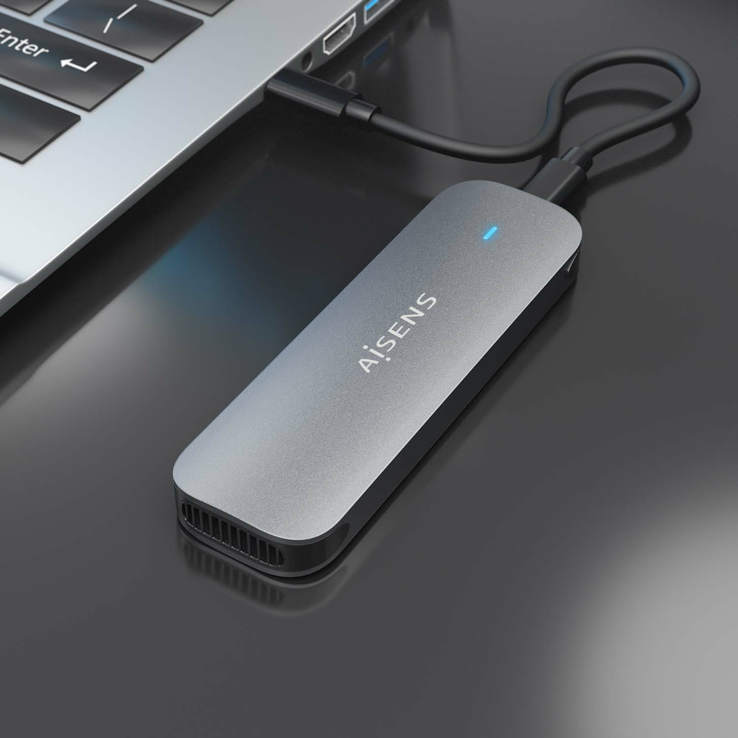 Un disco duro externo plateado conectado a una laptop. La unidad tiene la marca 'AISENS'. El fondo es una superficie oscura.