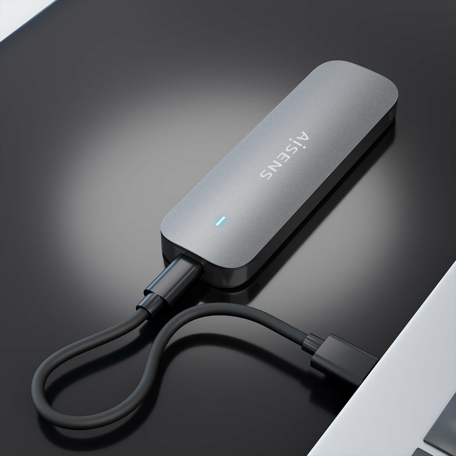 Un concentrador USB-C plateado AISENS conectado a una computadora portátil. Cable negro. El concentrador está sobre una superficie reflectante.