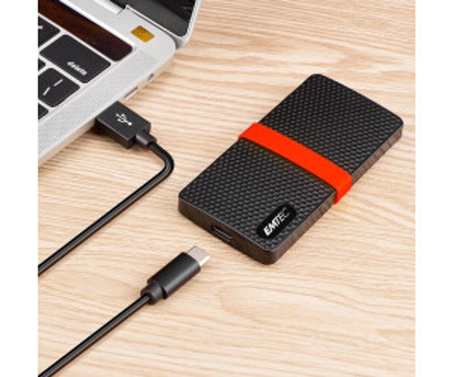 Czarny zewnętrzny dysk twardy z czerwonym paskiem podłączony do laptopa przez kabel USB. Drugi kabel USB-C leży na drewnianej powierzchni.