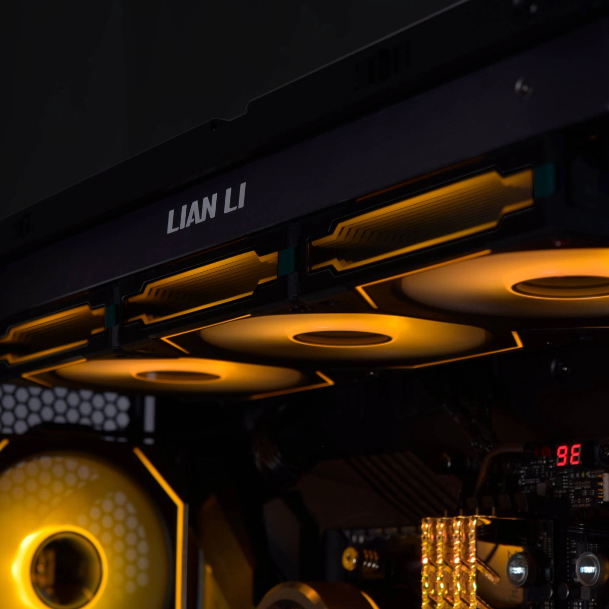 Composants PC avec ventilateurs éclairés, le logo Lian Li et un affichage numérique indiquant '98'. L'éclairage chaud et orange domine la scène.
