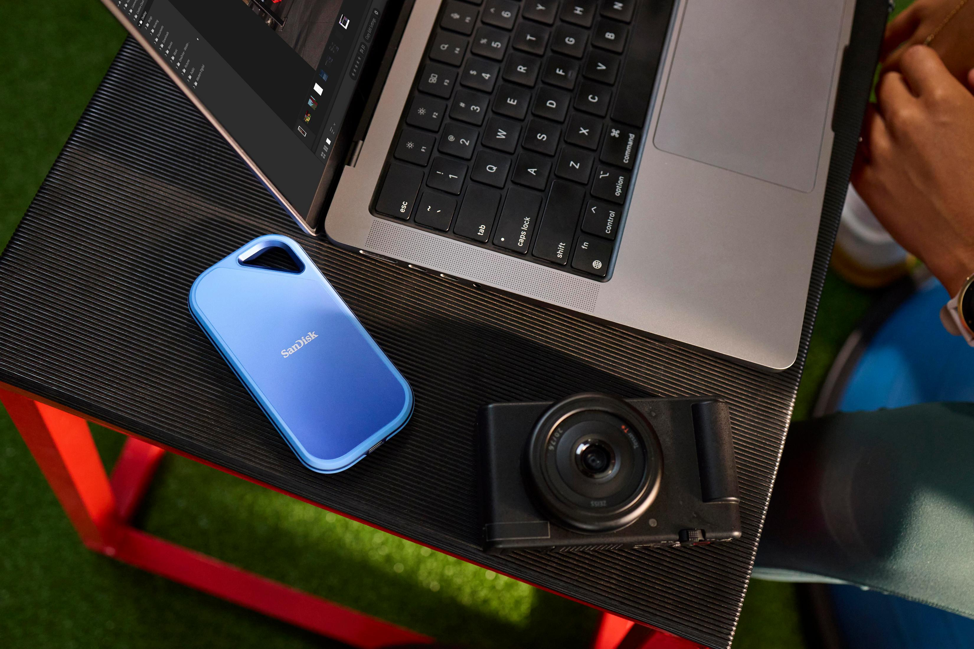 Laptop, aparat fotograficzny i niebieski dysk zewnętrzny na teksturowanej czarnej powierzchni. Zielona trawa i czerwona noga stołu w tle.