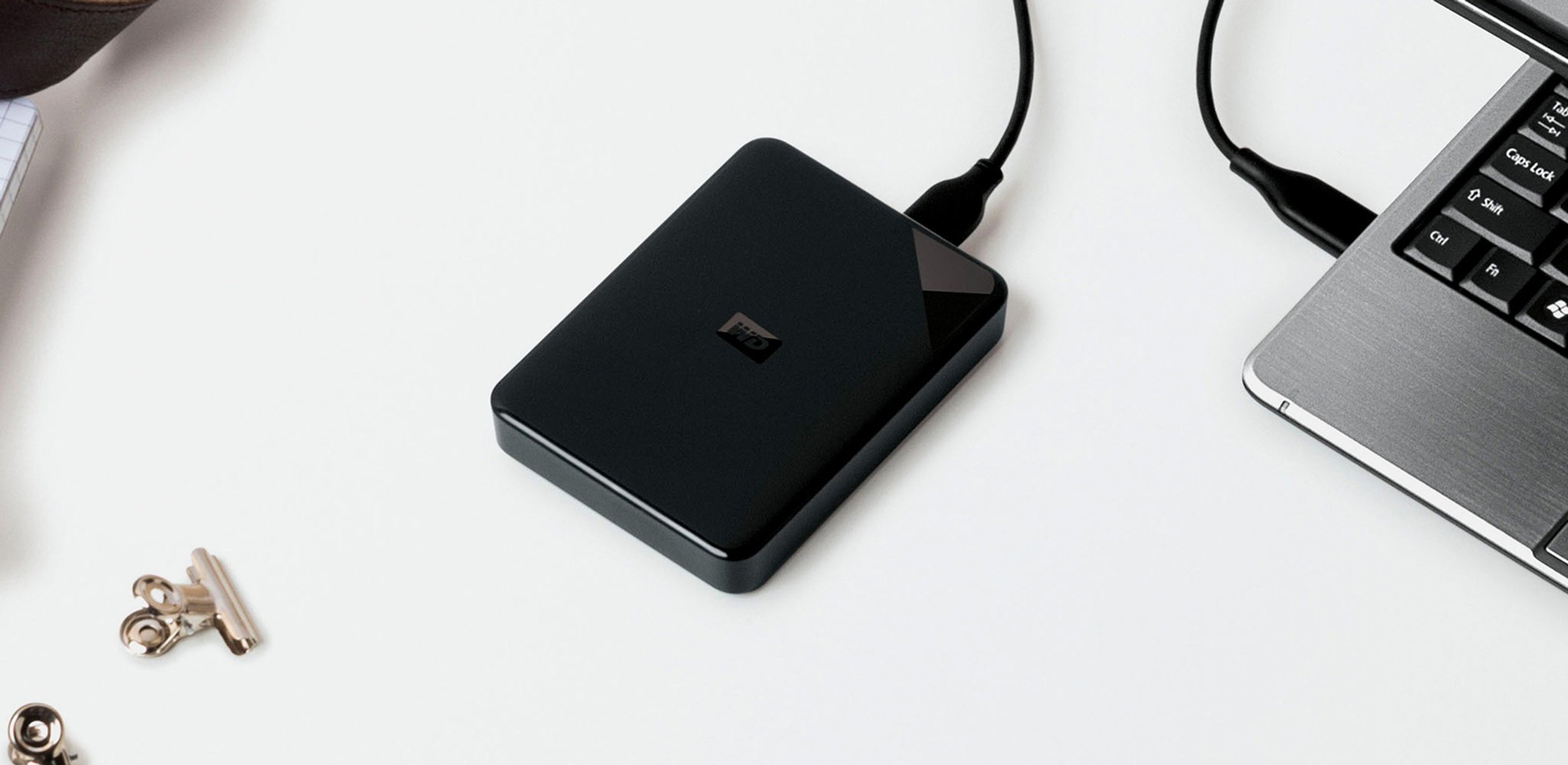 Czarny zewnętrzny dysk twardy podłączony do laptopa. Spinacz i notatnik na białej powierzchni.