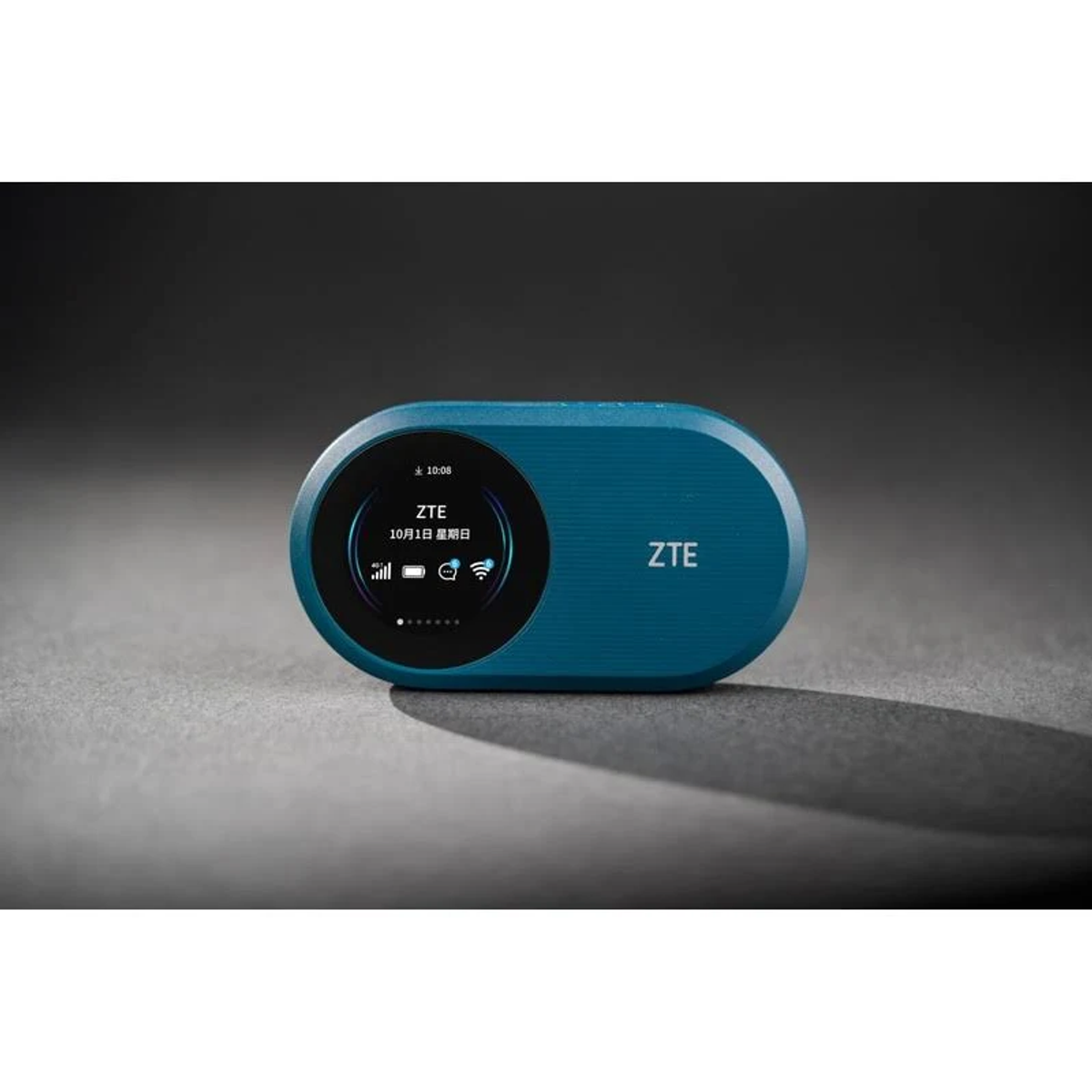 Turkusowe urządzenie ZTE na szarej powierzchni, wyświetlające godzinę i datę. Tło to szary gradient.