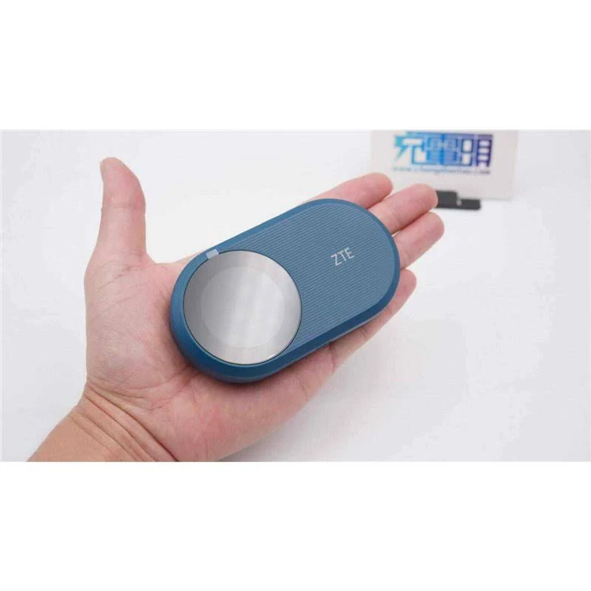 Dłoń trzyma niebieskie urządzenie ZTE. Jest owalne ze srebrnym okręgiem. Tło ma białe pudełko i chińskie znaki.