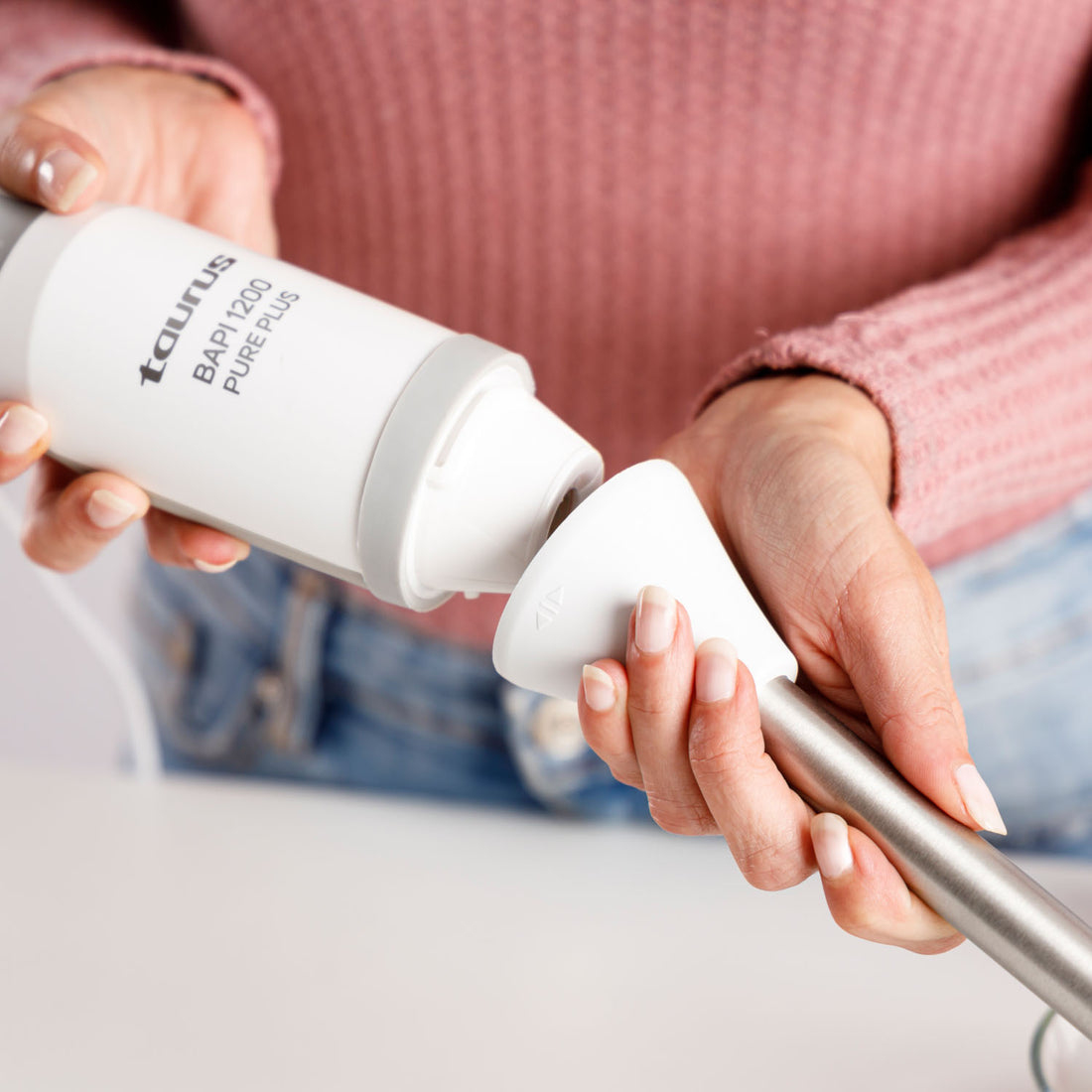 Osoba składa biały blender Taurus. Osoba ma na sobie różowy sweter i dżinsy. Części blendera są białe.