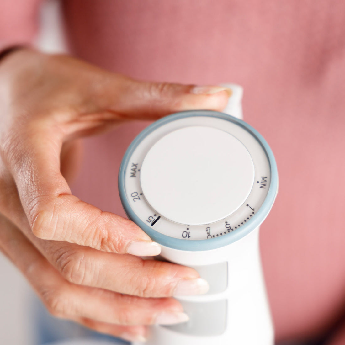 Ręka reguluje białe pokrętło z oznaczeniami temperatury. Znajduje się na białym urządzeniu na różowym tle. Pokrętło pokazuje MAX i MIN.