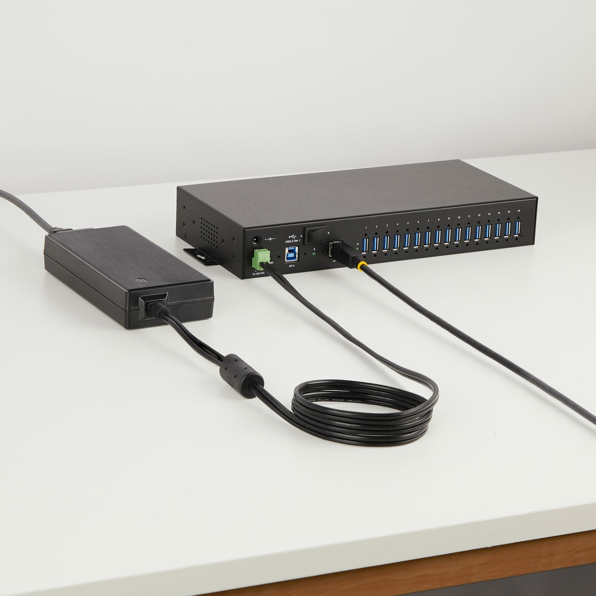 Ein schwarzer USB-Hub und Netzadapter auf einer weißen Oberfläche. Kabel verbinden sie. Der Hub hat mehrere USB-Anschlüsse.