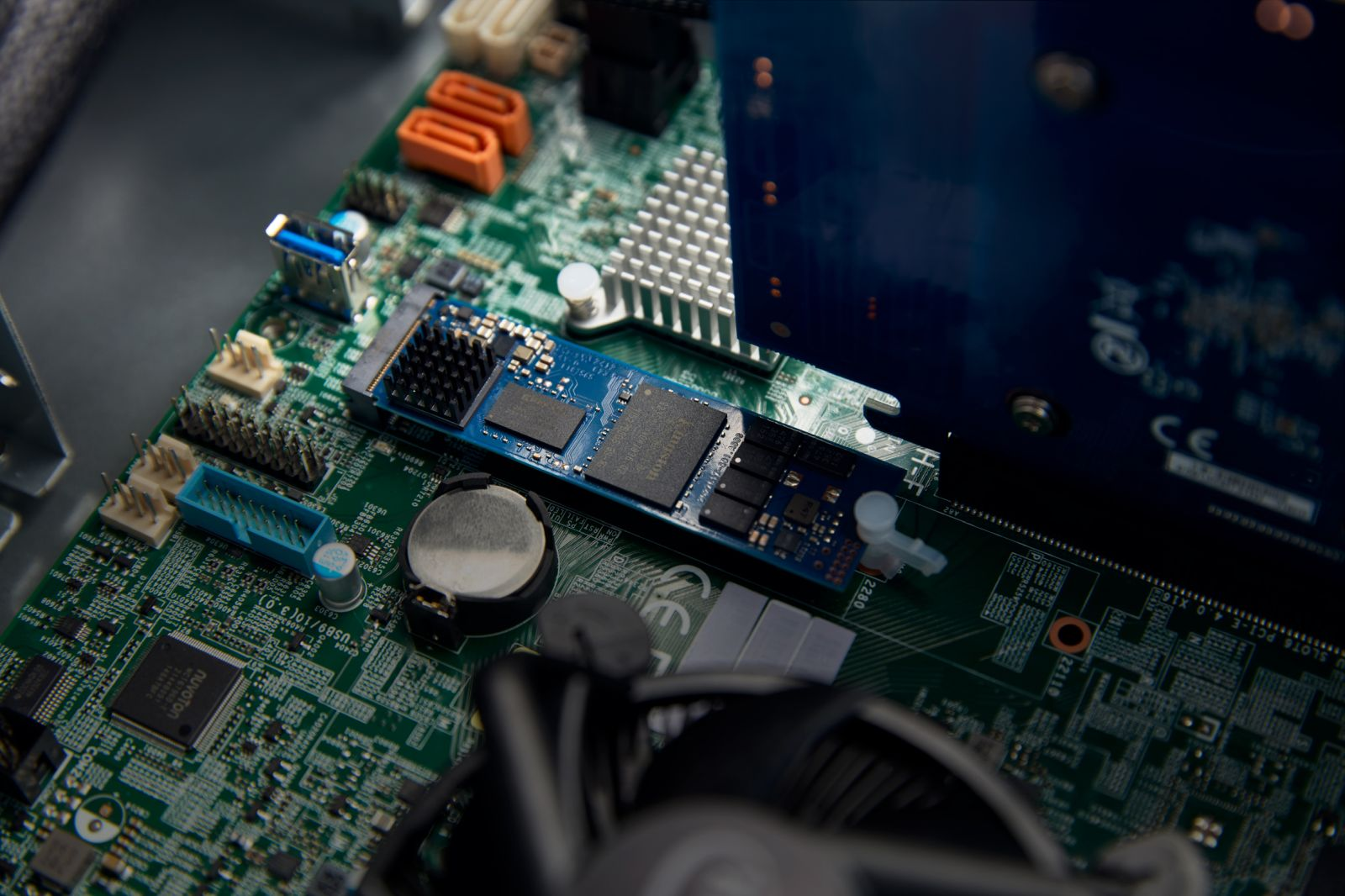 Zbliżenie płyty głównej komputera z zainstalowaną niebieską kartą SSD. Zielona płytka drukowana, pomarańczowe i niebieskie złącza.