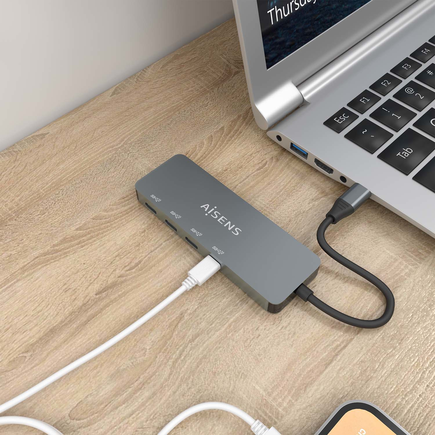 Un concentrador USB gris con cables blancos y negros está conectado a una computadora portátil sobre un escritorio de madera. Un teléfono está en la esquina.