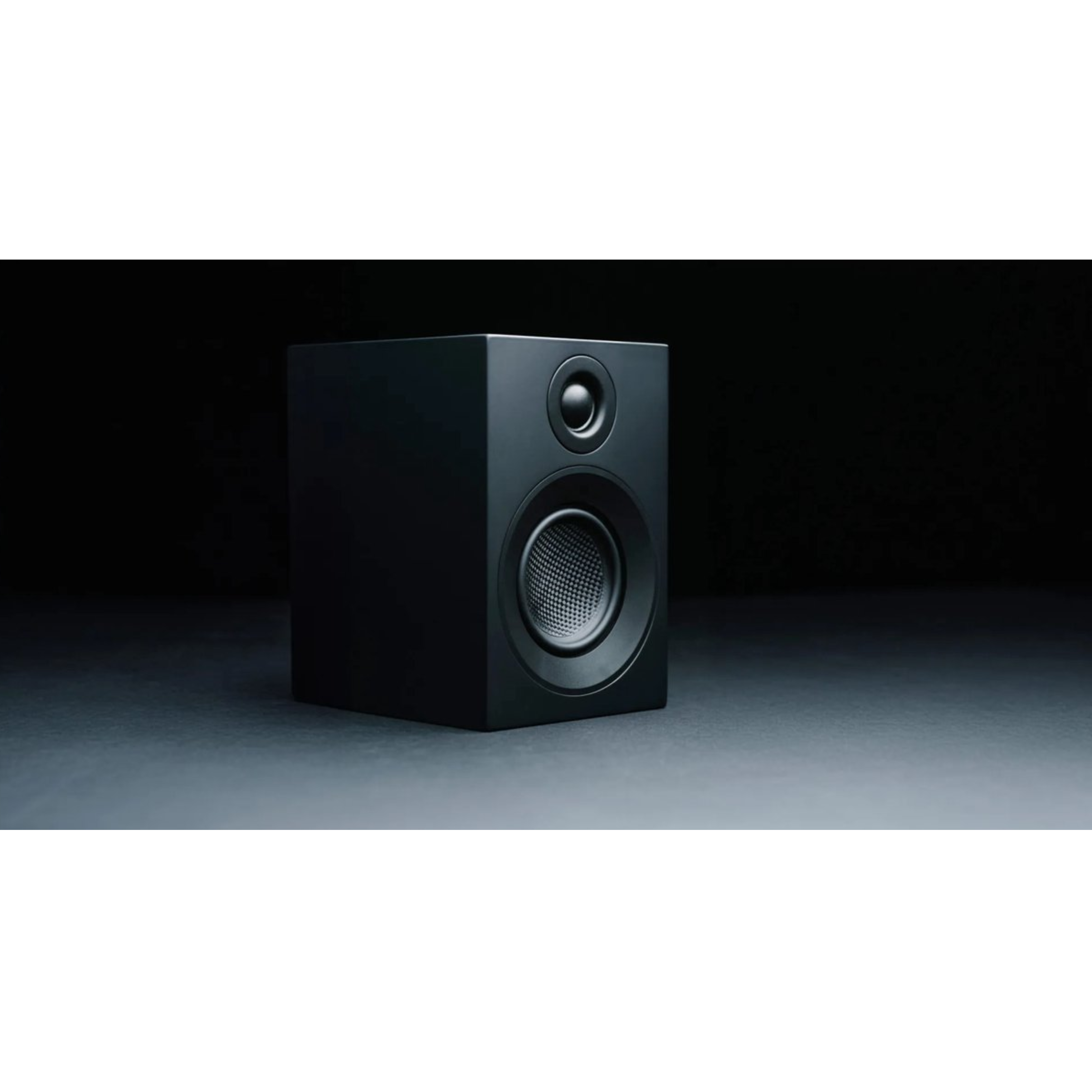 Czarny głośnik na ciemnej powierzchni, czarne tło. Posiada dwa okrągłe elementy głośnikowe.