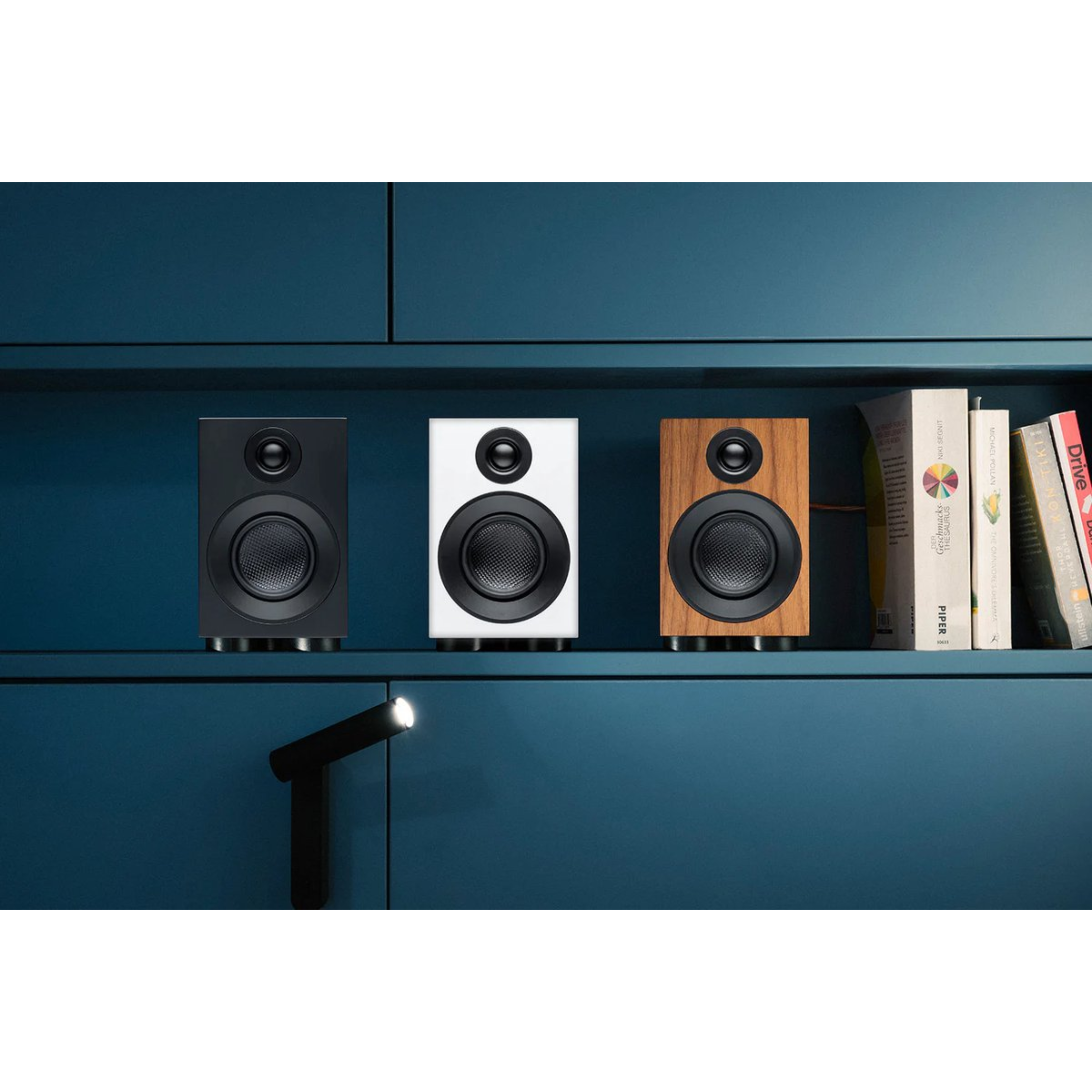 Trzy głośniki, czarne, białe i w kolorze drewna, stoją na półce. Książki po prawej stronie. Ciemnoniebieskie tło.