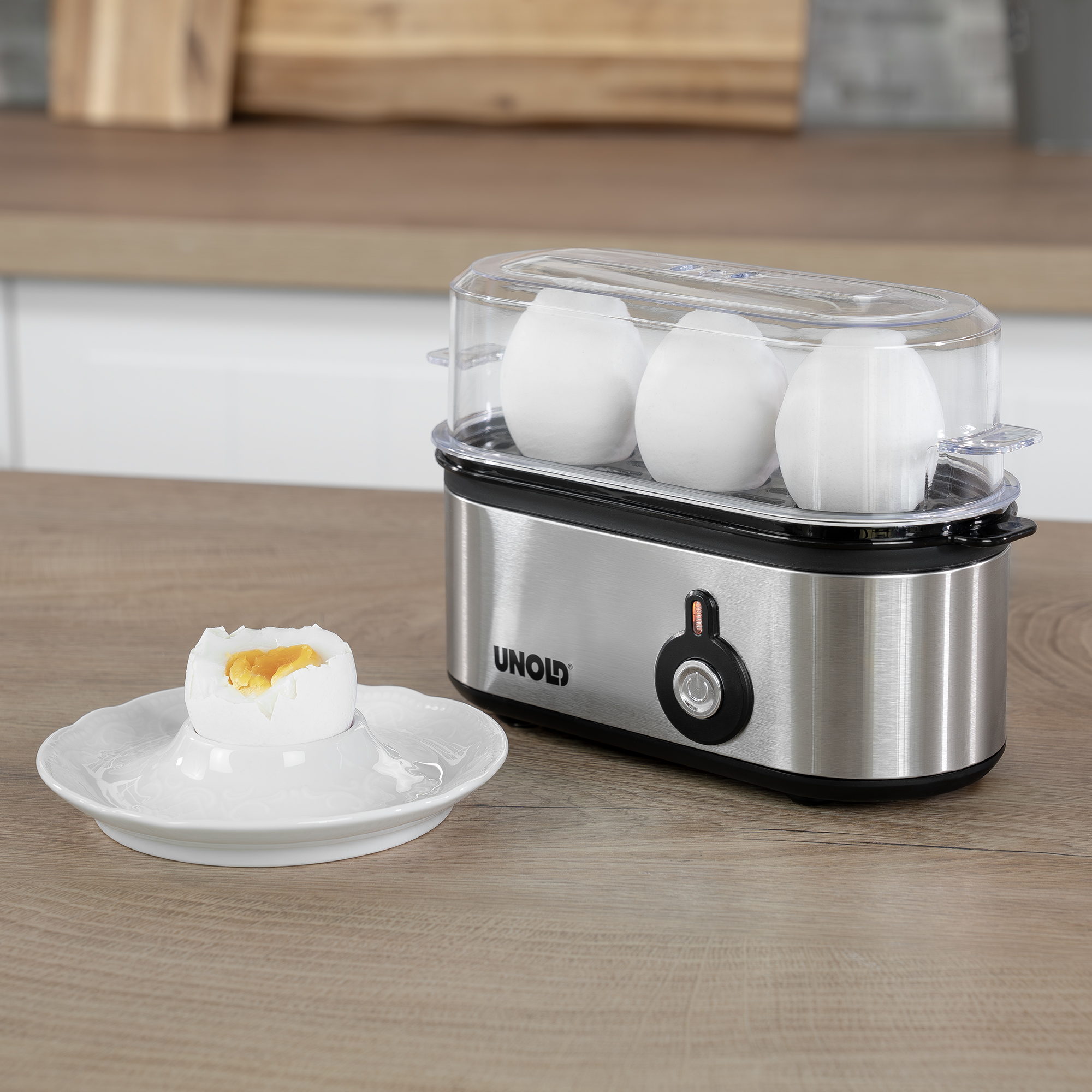 Urządzenie do gotowania jajek z jajkami w środku i jajkiem na talerzu. Jajko jest w połowie zjedzone, widoczny żółtek.