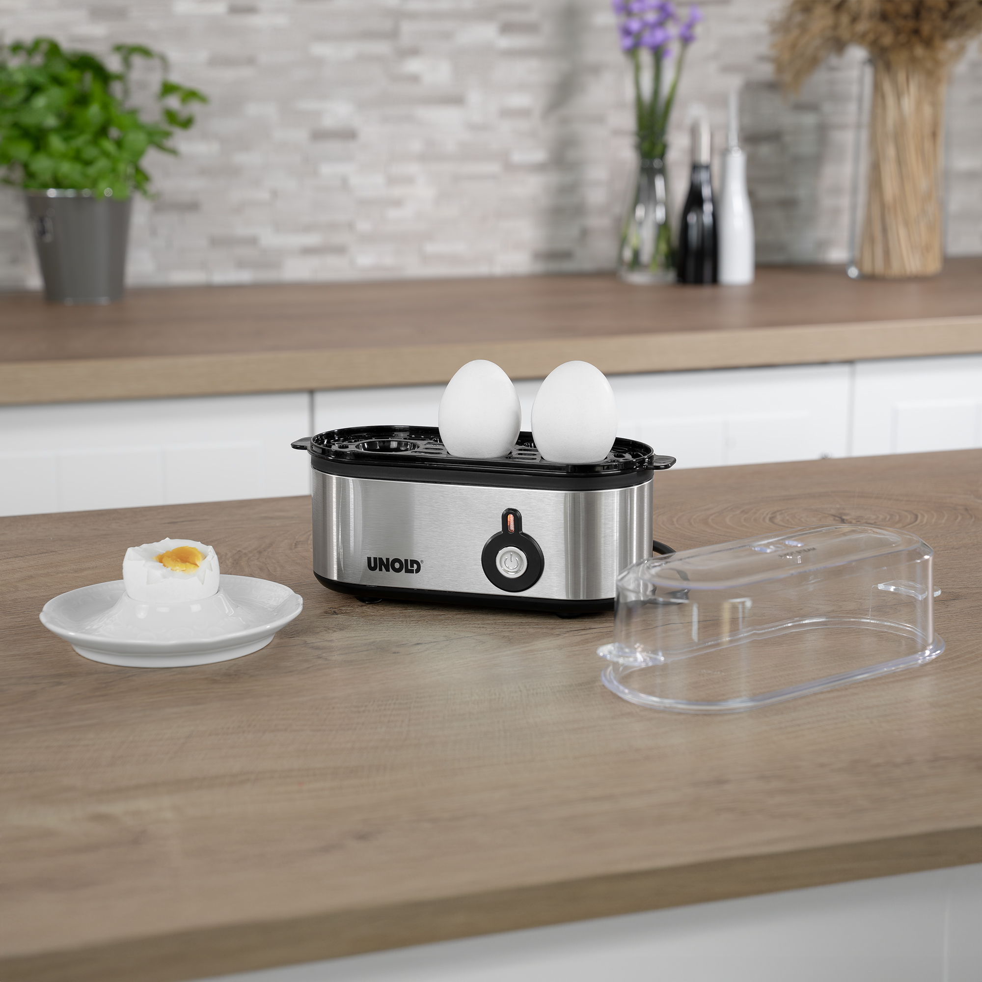 Gotowacz do jajek z dwoma jajkami, talerz z jajkiem i przezroczysta pokrywa na drewnianym blacie.