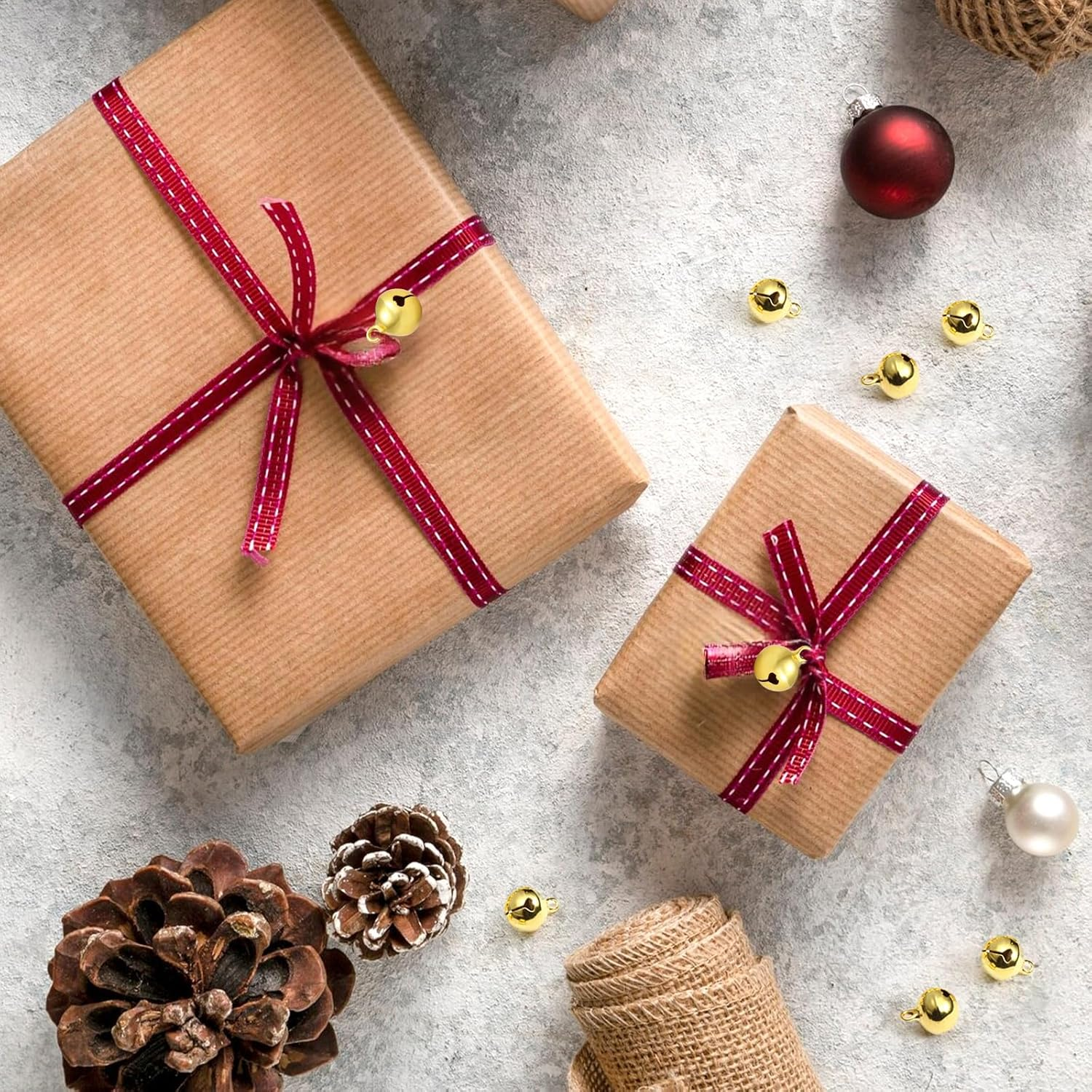 Zwei verpackte Geschenke mit roten Bändern und Glöckchen, Tannenzapfen, Ornamenten und Glöckchen auf hellem, grauem Untergrund.