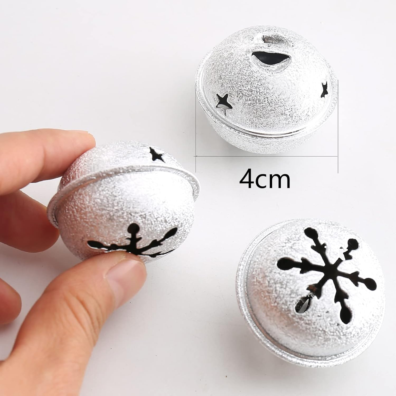 Drei silberne Glocken mit schwarzen Schneeflocken- und Sternausschnitten. Eine wird von Hand gehalten, 4 cm Messung sichtbar. Weißer Hintergrund.