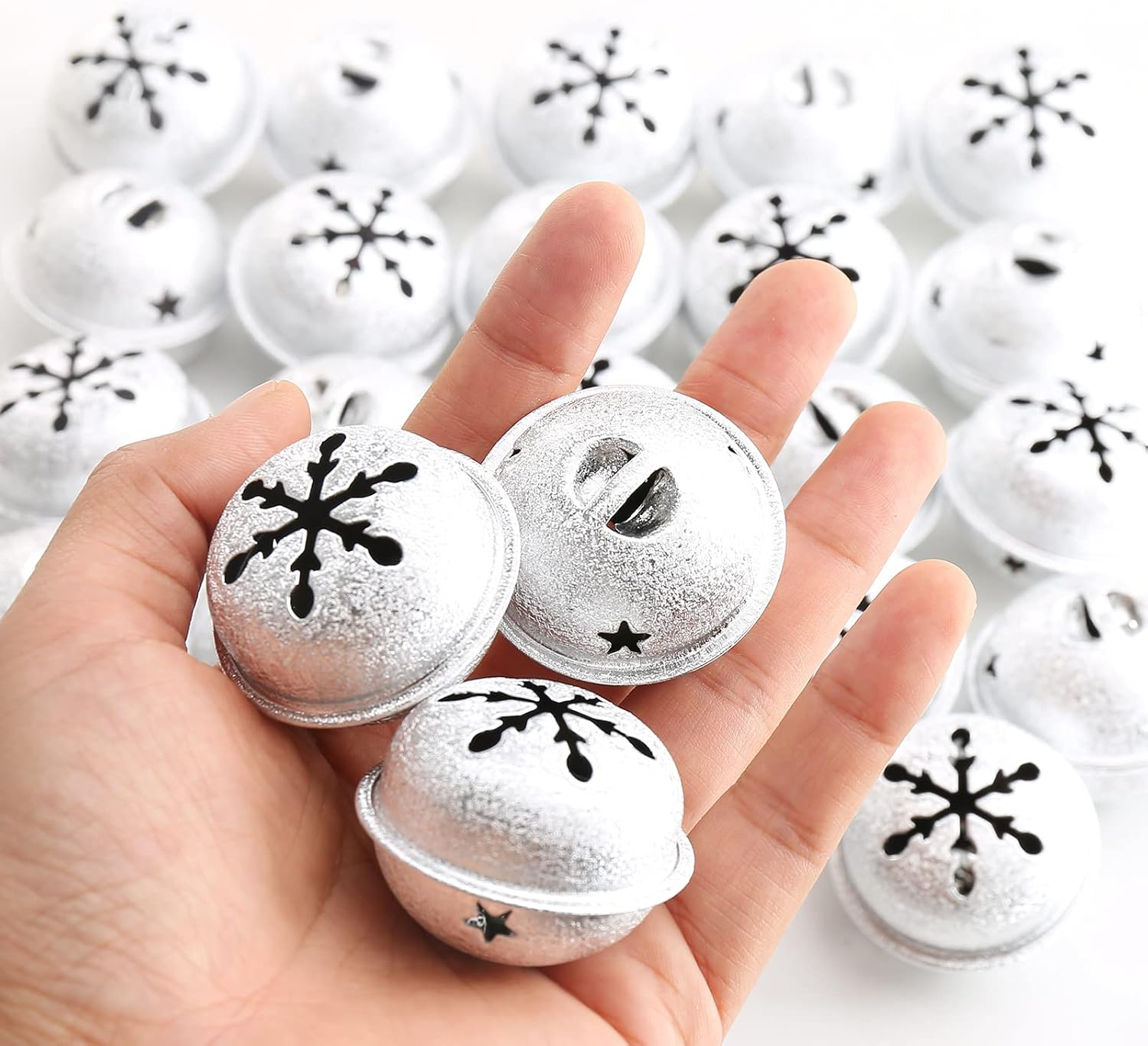 Eine Hand hält drei silberne Glöckchen mit Schneeflocken-Ausschnitten. Weitere Glocken sind im Hintergrund auf einer weißen Oberfläche.