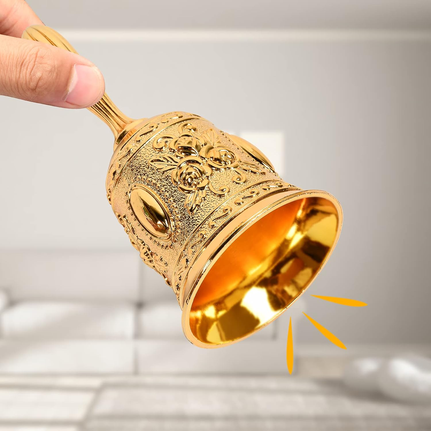 Eine Hand hält eine goldene Glocke mit floralen Mustern. Die Glocke ist geneigt, und orangefarbene Strahlen gehen von ihr aus.
