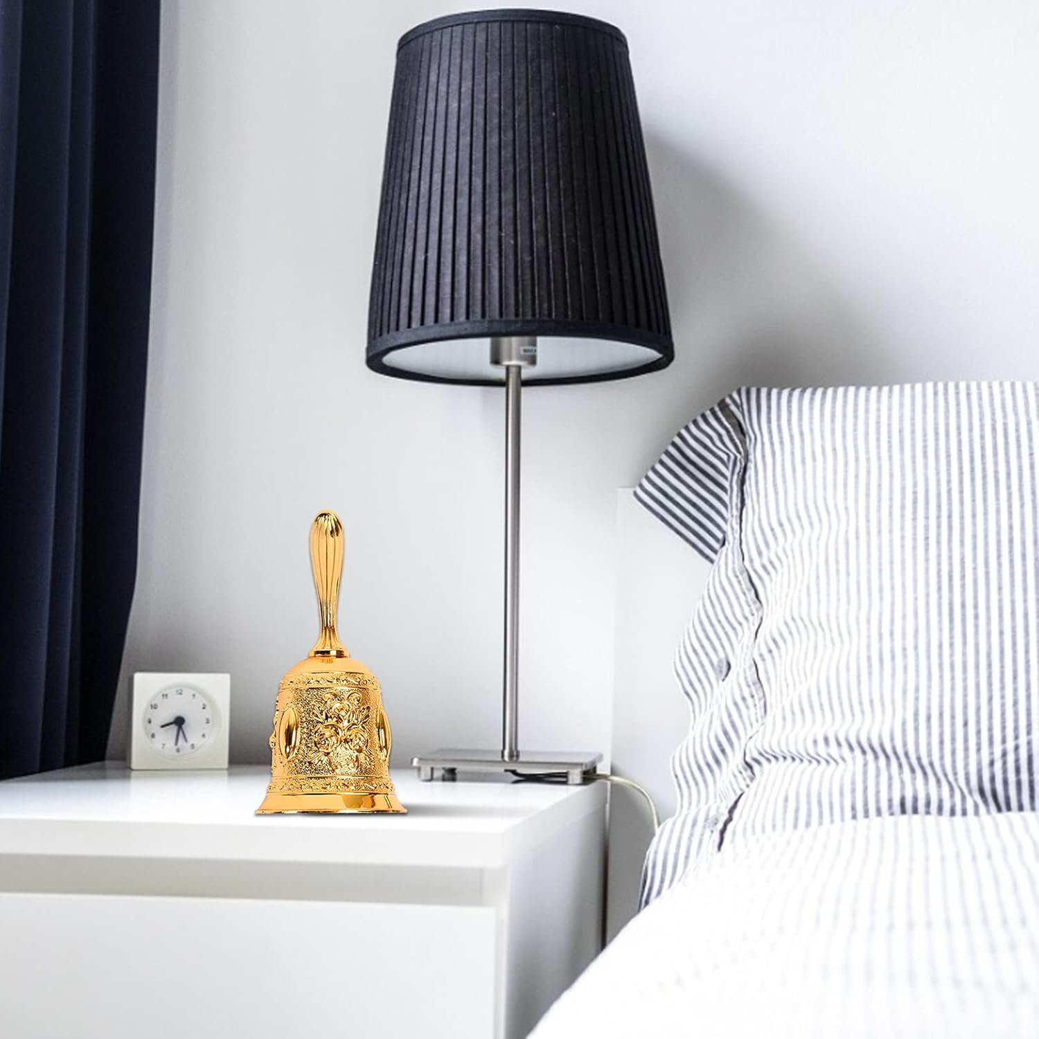 Eine goldene Glocke steht auf einem weißen Tisch neben einer Lampe und einer kleinen Uhr. Im Hintergrund befindet sich ein Bett mit gestreifter Bettwäsche.