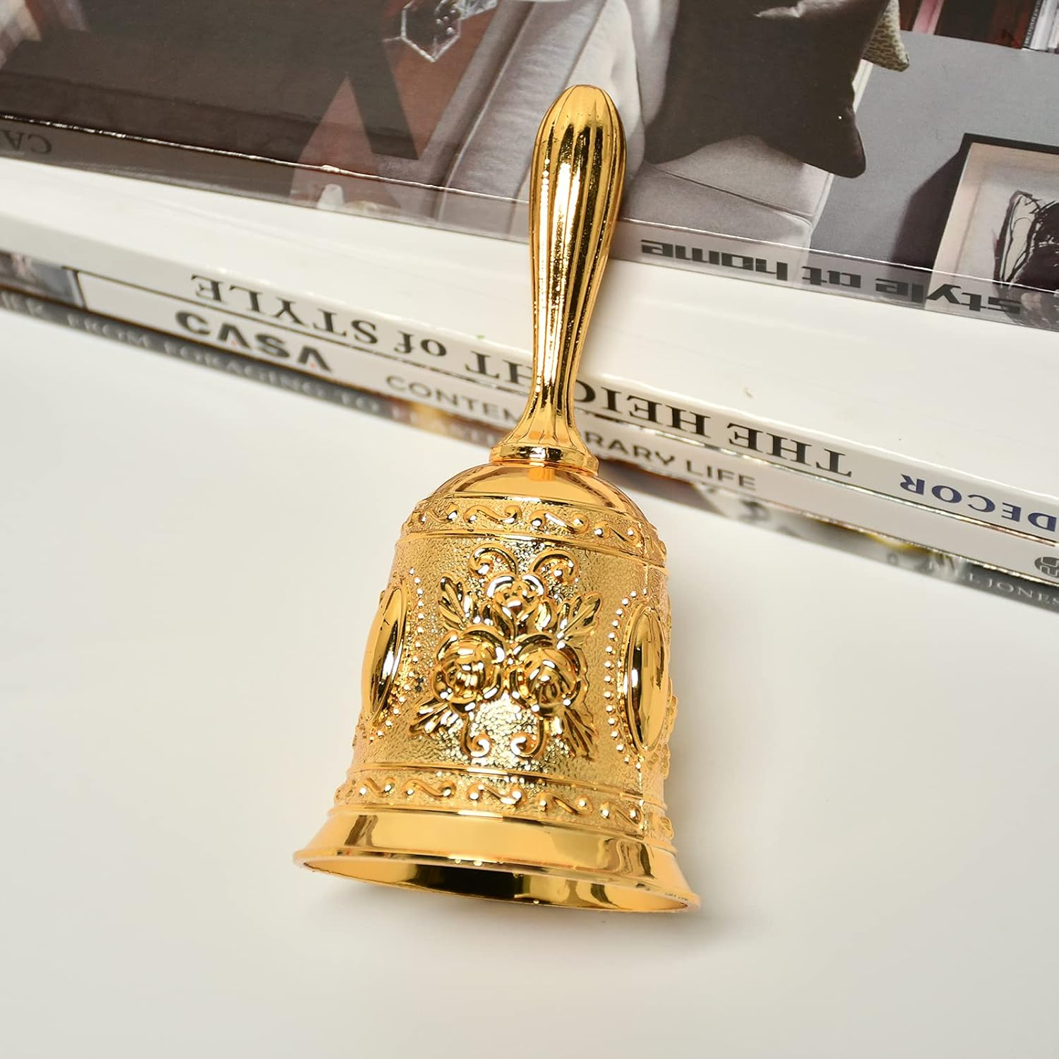 Eine goldene Glocke mit einem verzierten Blumendesign auf einer weißen Oberfläche mit Büchern im Hintergrund.