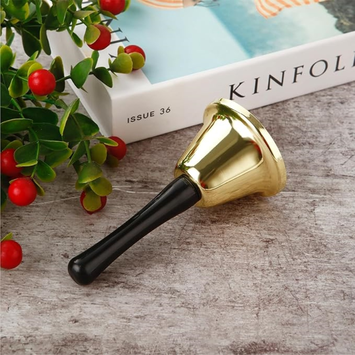 Eine goldene Glocke mit schwarzem Griff auf einer Holzoberfläche. Daneben grüne Blätter mit roten Beeren und ein Buch.