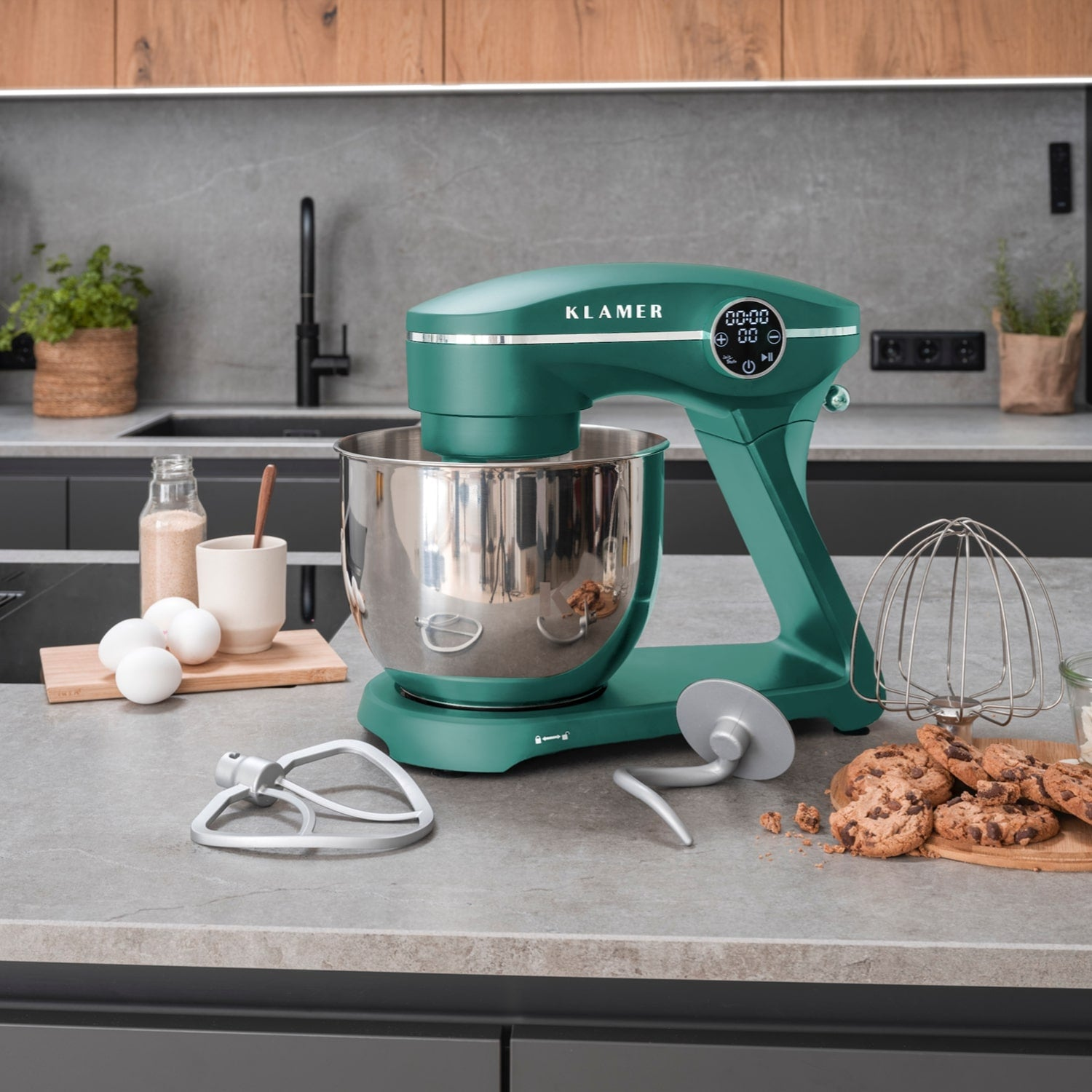 Ein grüner Küchenmixer mit Edelstahlschüssel steht auf einer Theke. Kekse, Eier und andere Gegenstände sind in der Nähe.