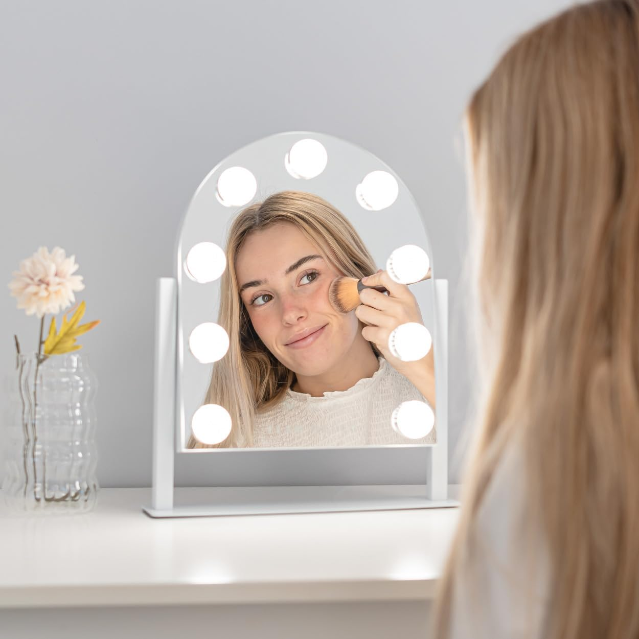 Una mujer se aplica maquillaje con un pincel, reflejada en un espejo con luces. Un jarrón con flores está sobre una mesa.