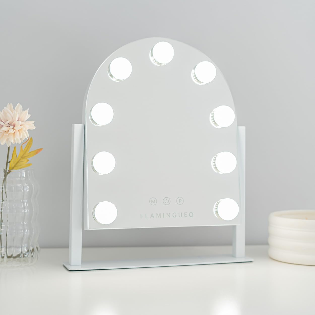 Un espejo de maquillaje blanco con luces, un jarrón con flores y una vela están sobre una superficie blanca contra una pared gris clara.