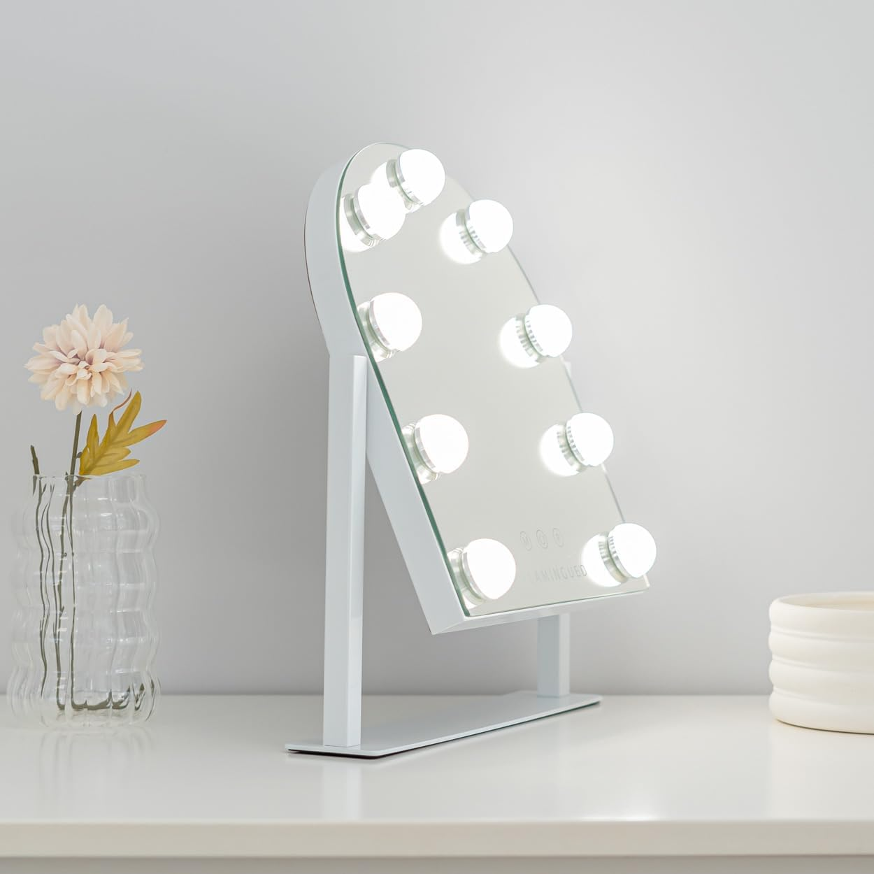 Un espejo de tocador blanco con luces está sobre una mesa blanca. Una flor y un jarrón a la izquierda. Un recipiente blanco a la derecha.