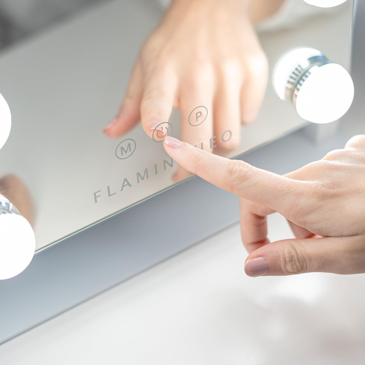 Una mano toca un espejo con bombillas, presionando un botón de encendido. La palabra 'Flamingo' es visible. El espejo refleja una mano.