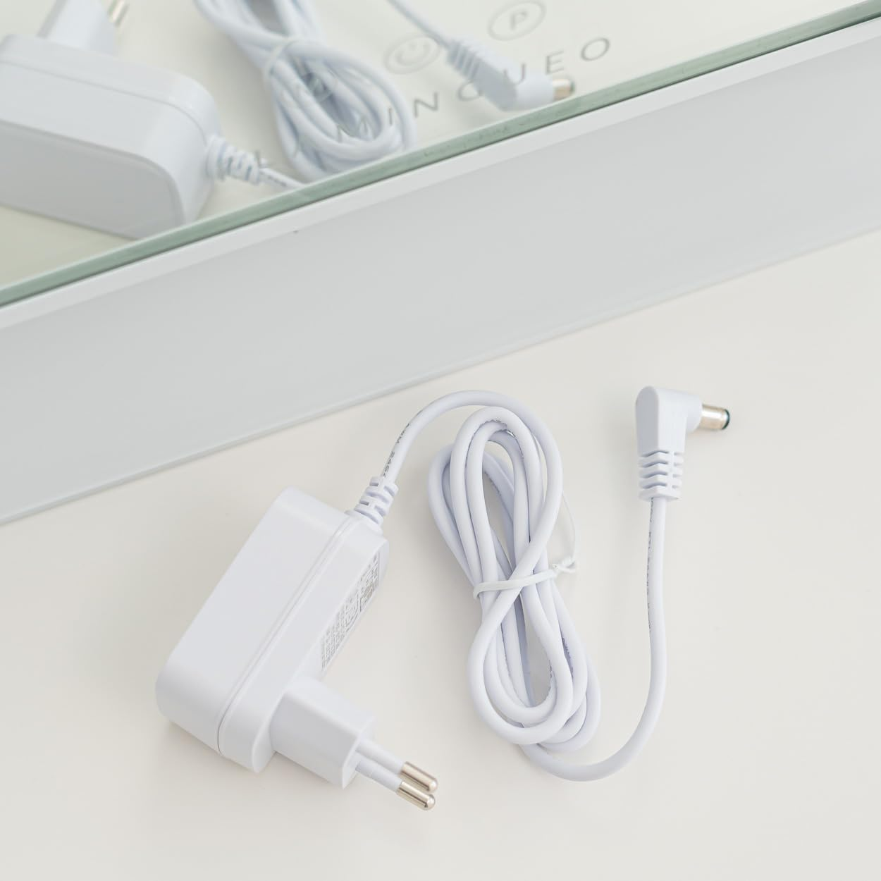 Un adaptador de corriente y un cable blancos sobre una superficie blanca. Un espejo y parte de un dispositivo al fondo. Escena bien iluminada.