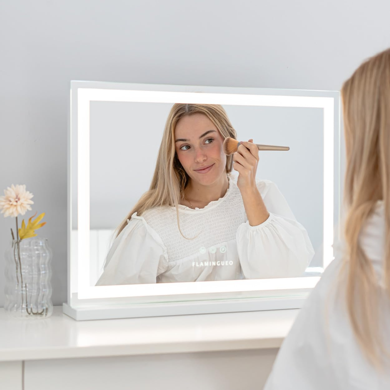 Mujer maquillándose en un espejo iluminado. El espejo refleja su cara, un pincel y su camisa blanca. Flores a su lado.
