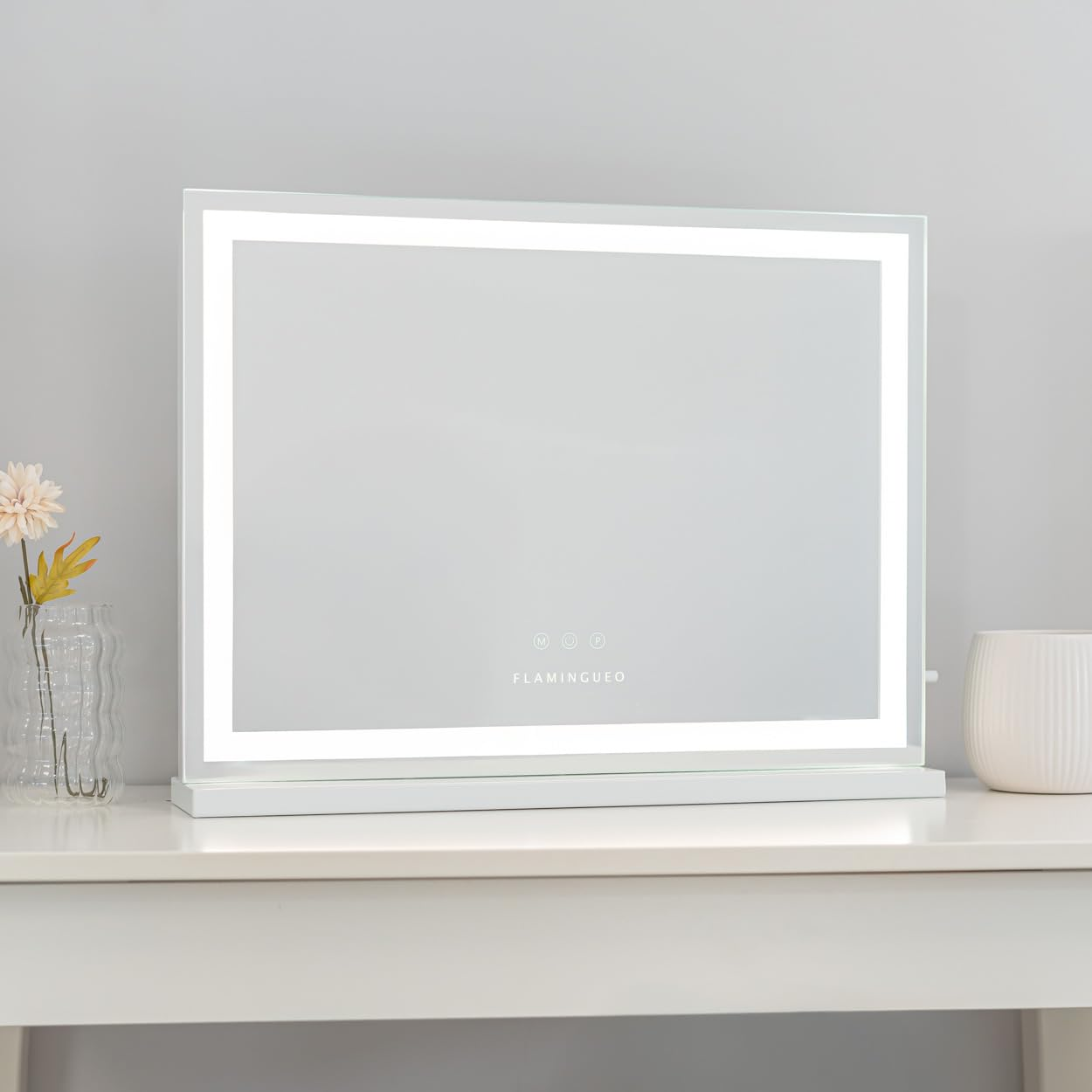Un espejo de maquillaje LED blanco está sobre una mesa blanca. Un jarrón con flores a la izquierda y una maceta blanca a la derecha.