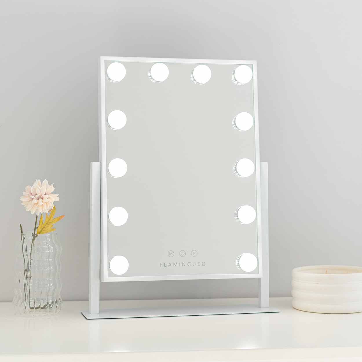 Un espejo de maquillaje blanco con luces está sobre una superficie blanca. Una flor en un jarrón y una vela están al lado.