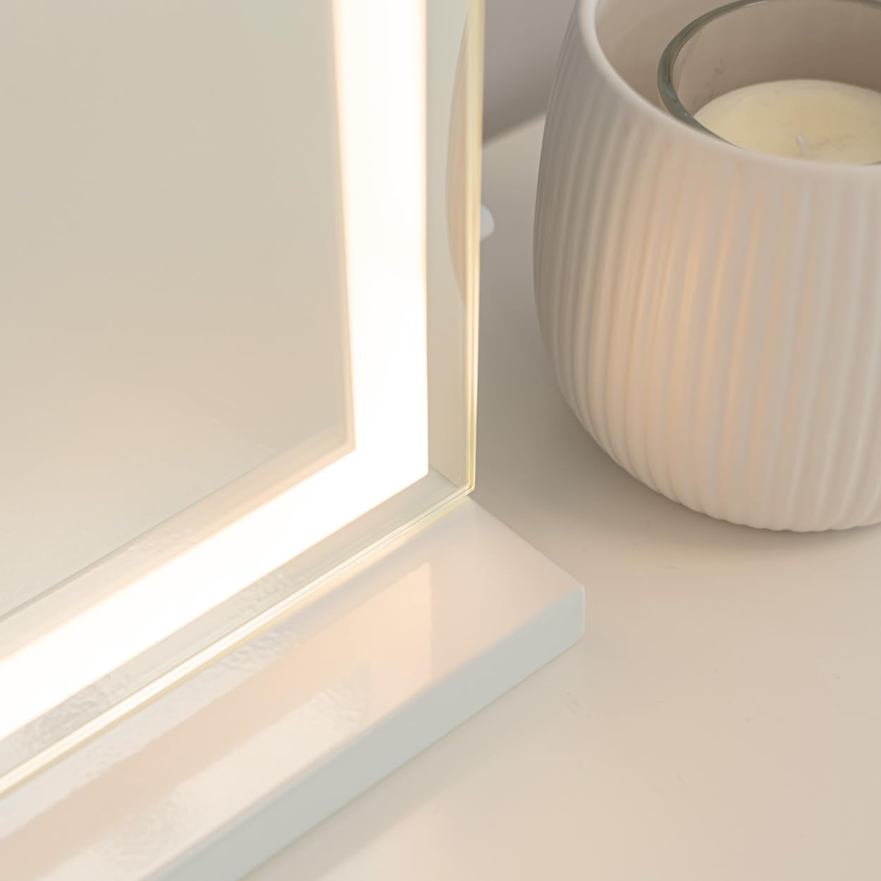 Un espejo iluminado y un portavelas blanco y acanalado sobre una superficie blanca. El espejo es rectangular e iluminado.