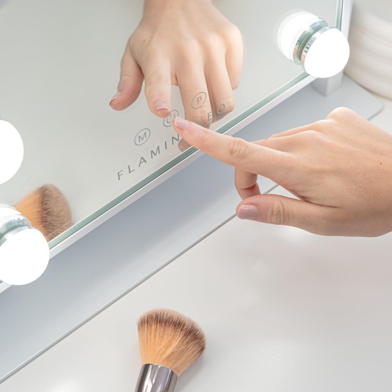 Una mano toca un espejo con luces. Hay una brocha de maquillaje en la parte inferior. El espejo tiene botones de encendido y modo.